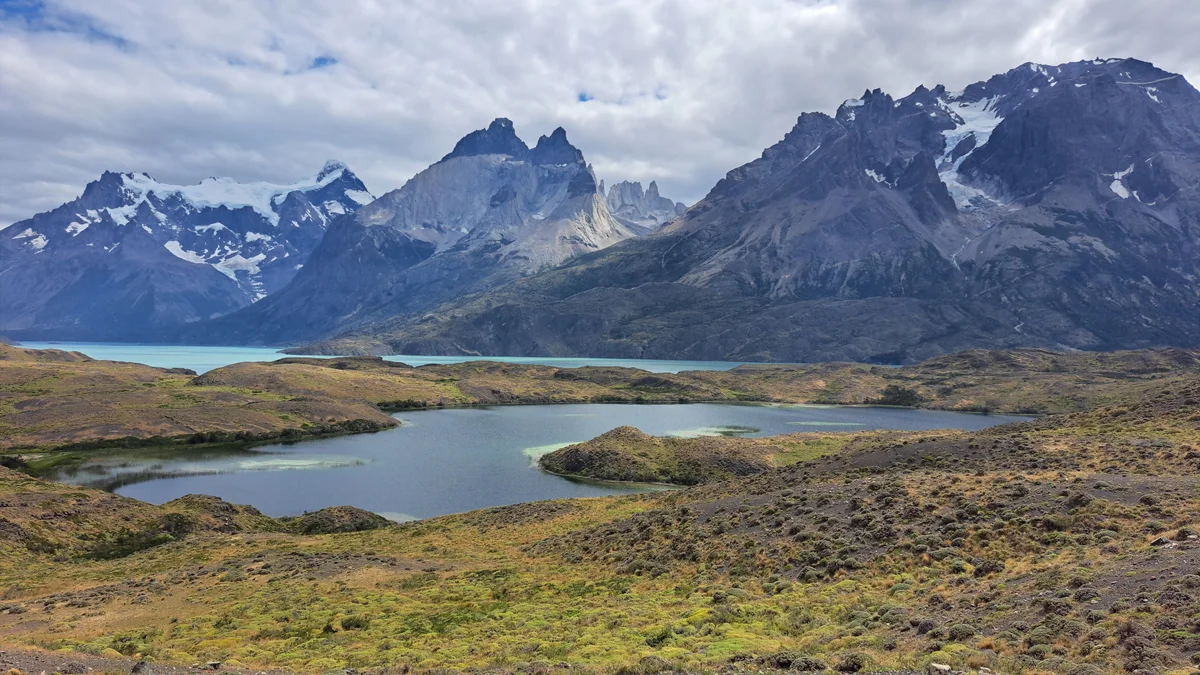 Paisaje de montaña con picos nevados, lagos de agua azul y vegetación de estepa bajo un cielo nublado.