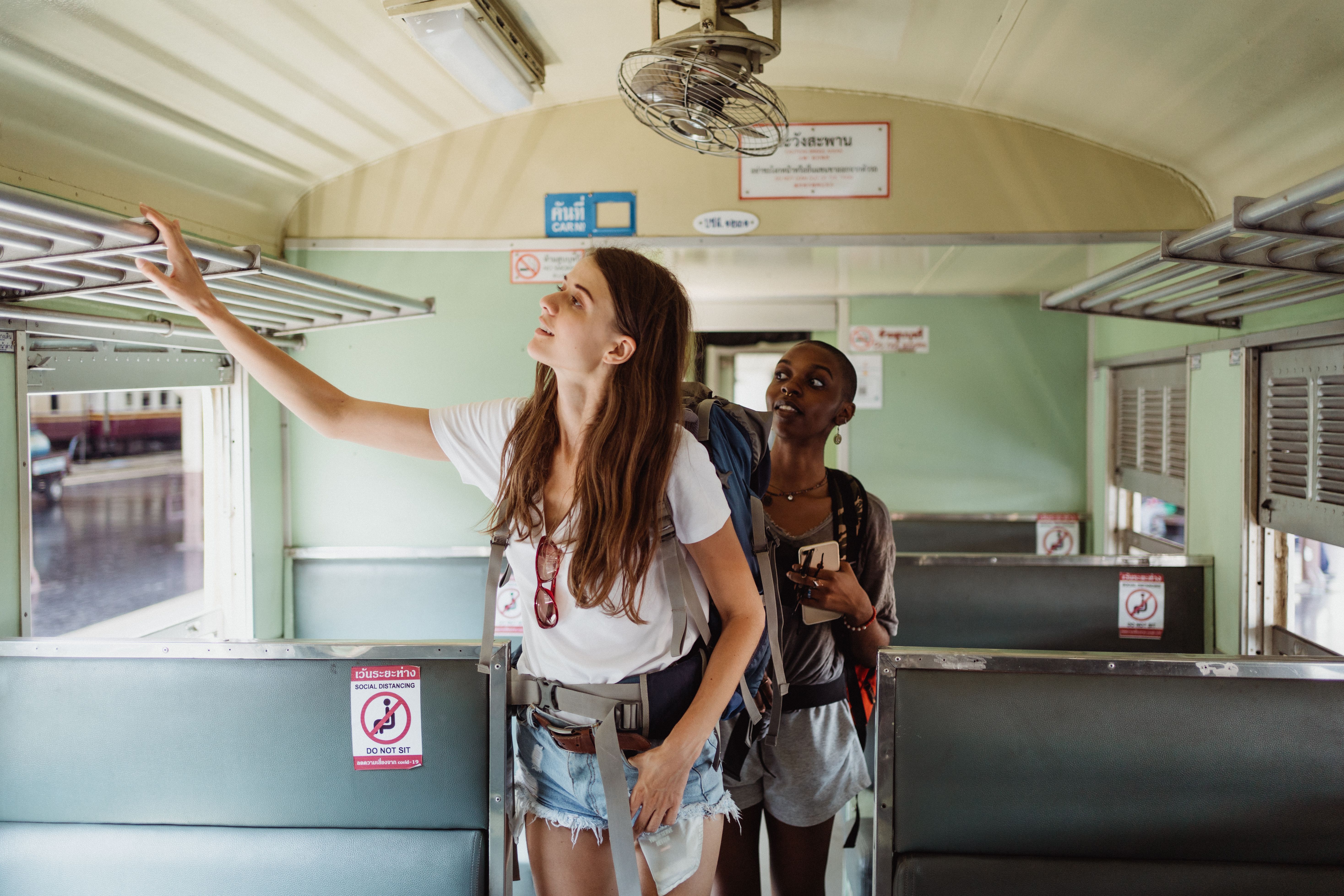 Dos mujeres jóvenes con mochilas dentro de un vagón de tren tailandés, una alcanzando un estante superior.