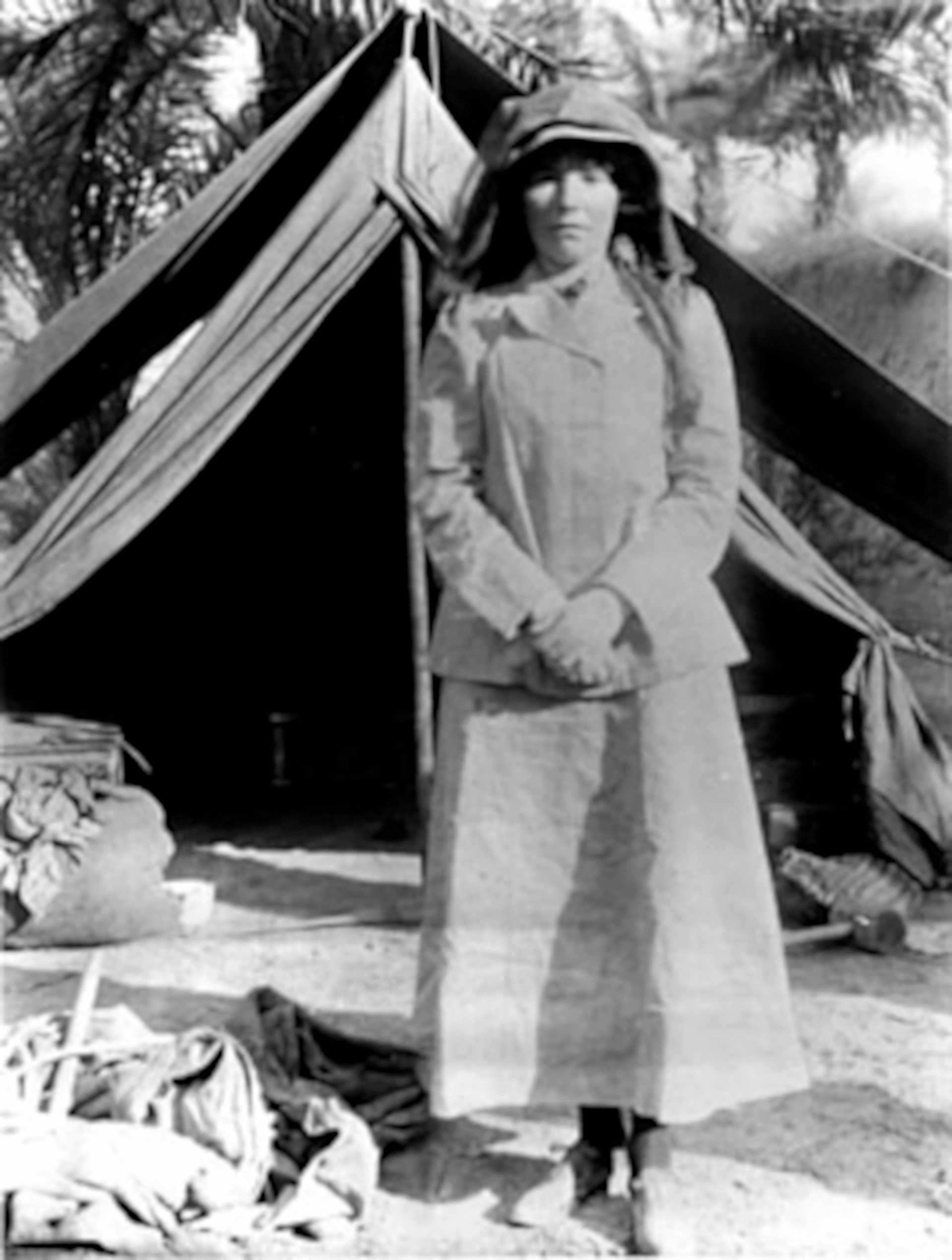 Retrato histórico de Gertrude Bell de pie frente a una tienda de campaña en un campamento desértico, con palmeras al fondo