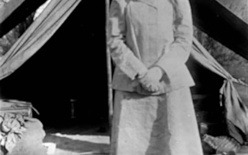 Retrato histórico de Gertrude Bell de pie frente a una tienda de campaña en un campamento desértico, con palmeras al fondo