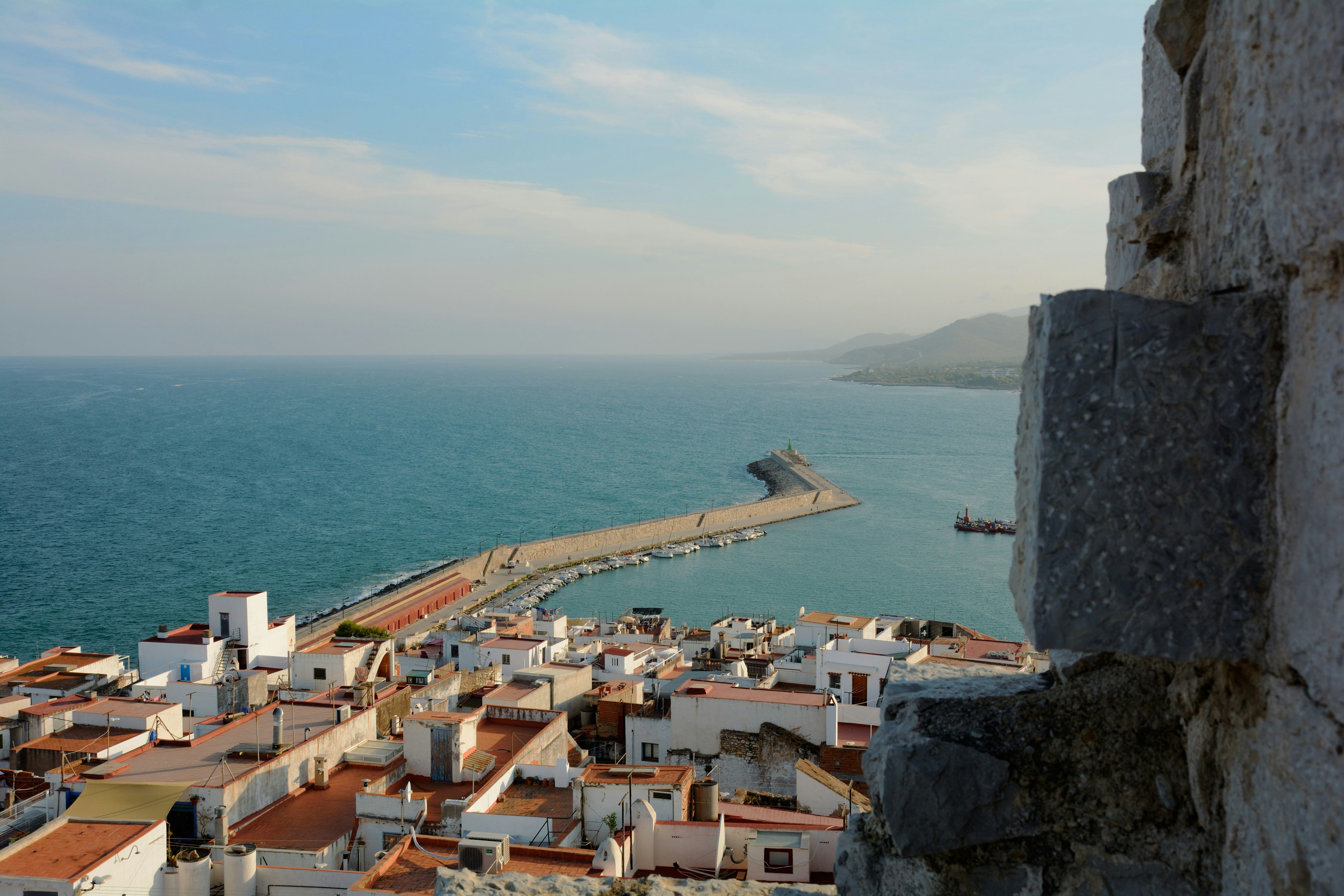 Vista aérea de un pueblo costero con casas blancas y tejados naranjas, un puerto y el mar azul.