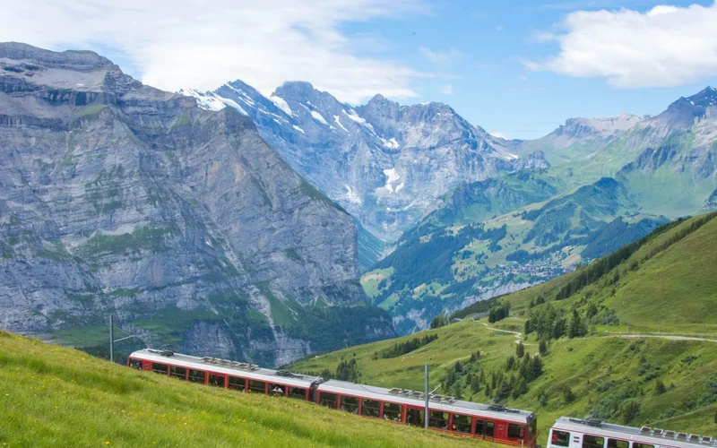 Tren rojo y blanco ascendiendo una ladera verde con montañas nevadas al fondo bajo cielo azul.
