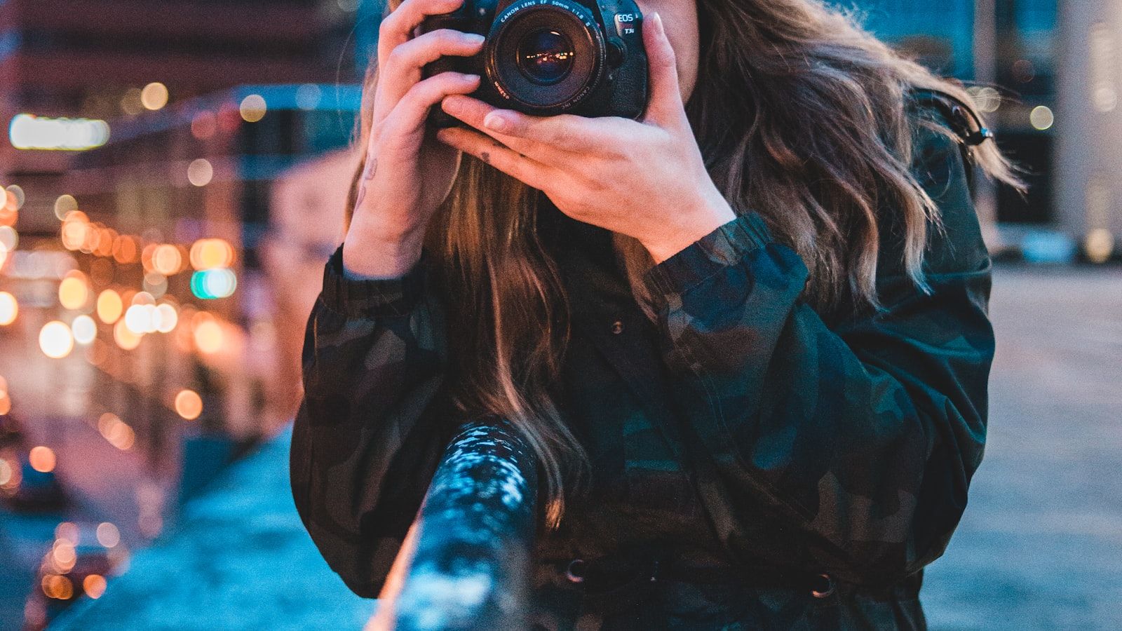 A night street photographer shooting handheld with a Canon EOS 77D and 50mm f/1.8 STM lens, foreground hands cupping the camera in front of their face — bokeh streetlights frame the shot