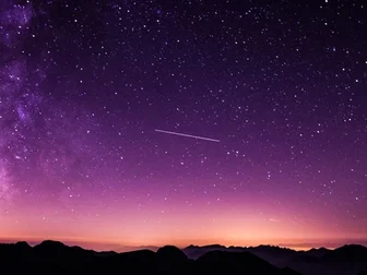 A purple and orange Milky Way galaxy stretches across the night sky above a silhouetted mountain ridge, with a shooting star crossing the frame.