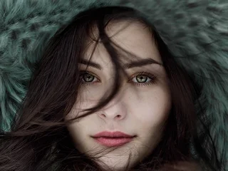 Dark moody close-up portrait of a young woman with dark hair, green eyes, partially obscured by windblown hair and a teal fur hood - atmospheric reference for a Photoshop composite workflow cover