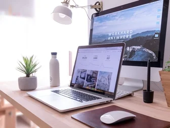 A clean dual-monitor desk workspace with a laptop showing a photo editing dashboard, paired with a desktop display showing a mountain landscape, plant and desk lamp on the side.