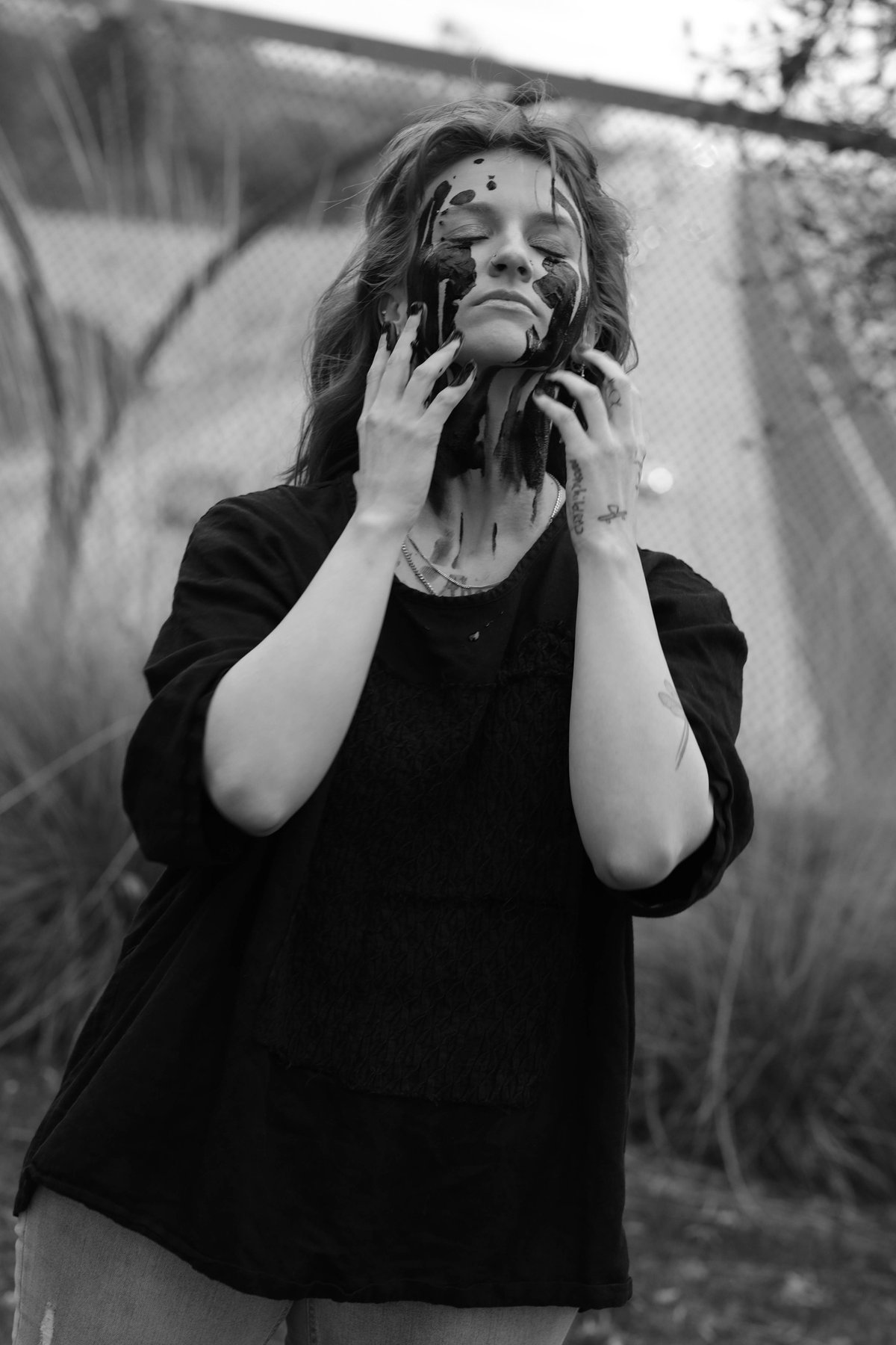 A woman stands with her eyes closed, hands at her face and neck, black paint running from her eyes.
