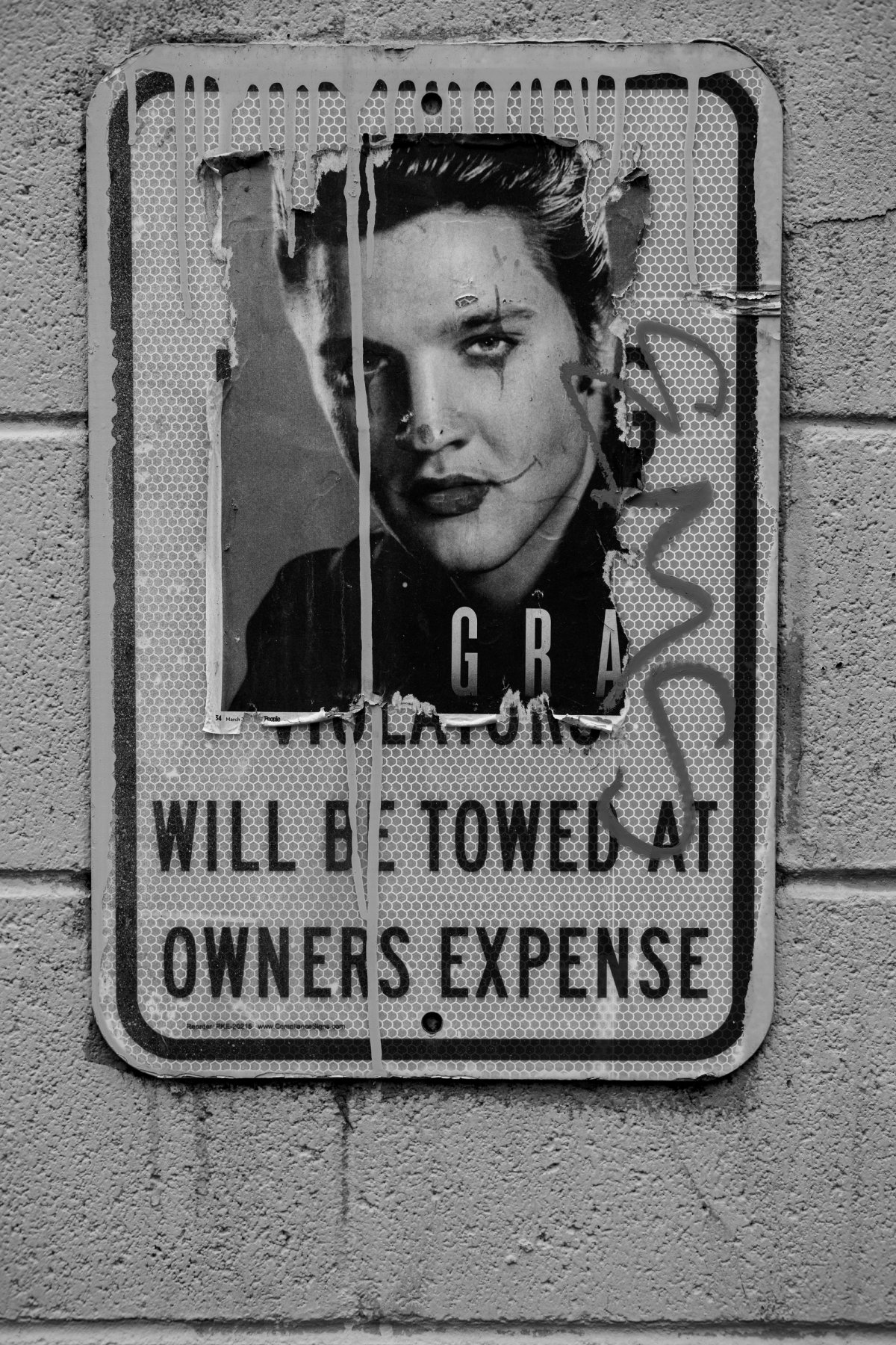A 'will be towed at owner's expense' sign with a torn poster of a young man pasted over it.
