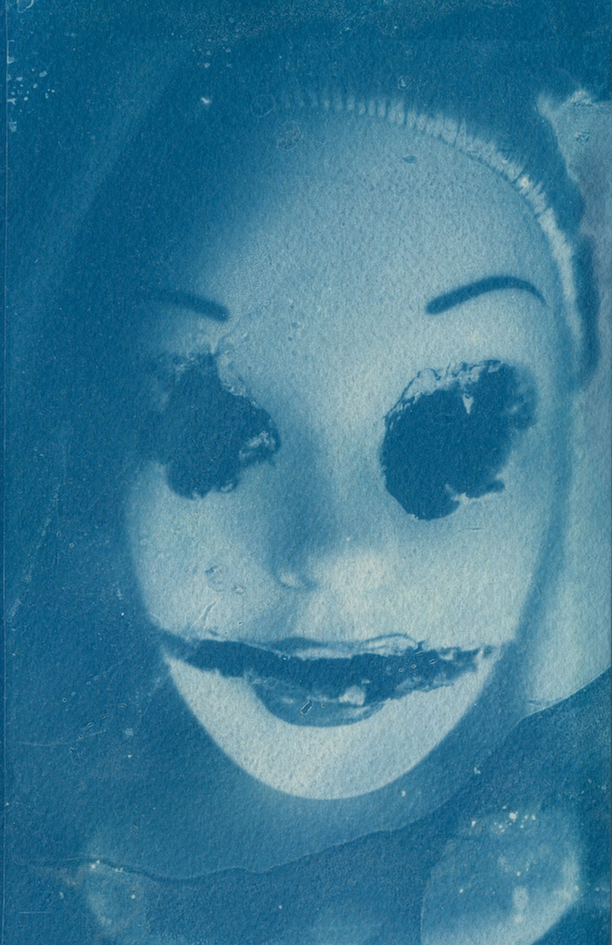 A cyanotype of a doll's face with blotted-out eyes and a smeared smile.