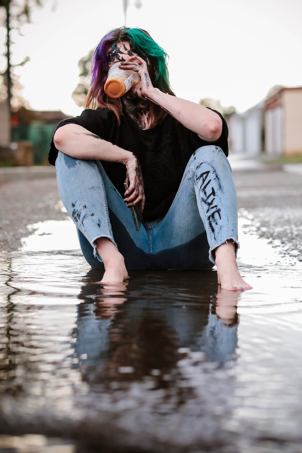 A woman with purple and green hair sits barefoot in a puddle on a road, drinking from a jar, 'ALIVE' painted on her jeans.