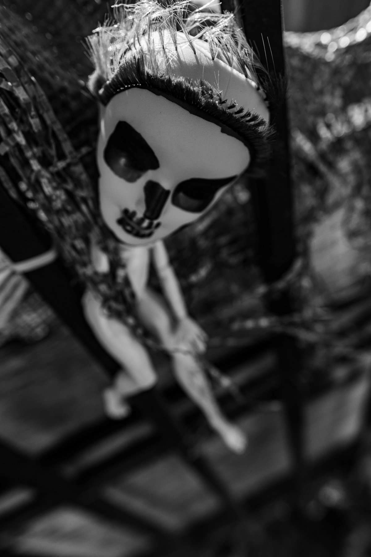 A doll with a white skull face and tufted hair, bound in tinsel behind bars.