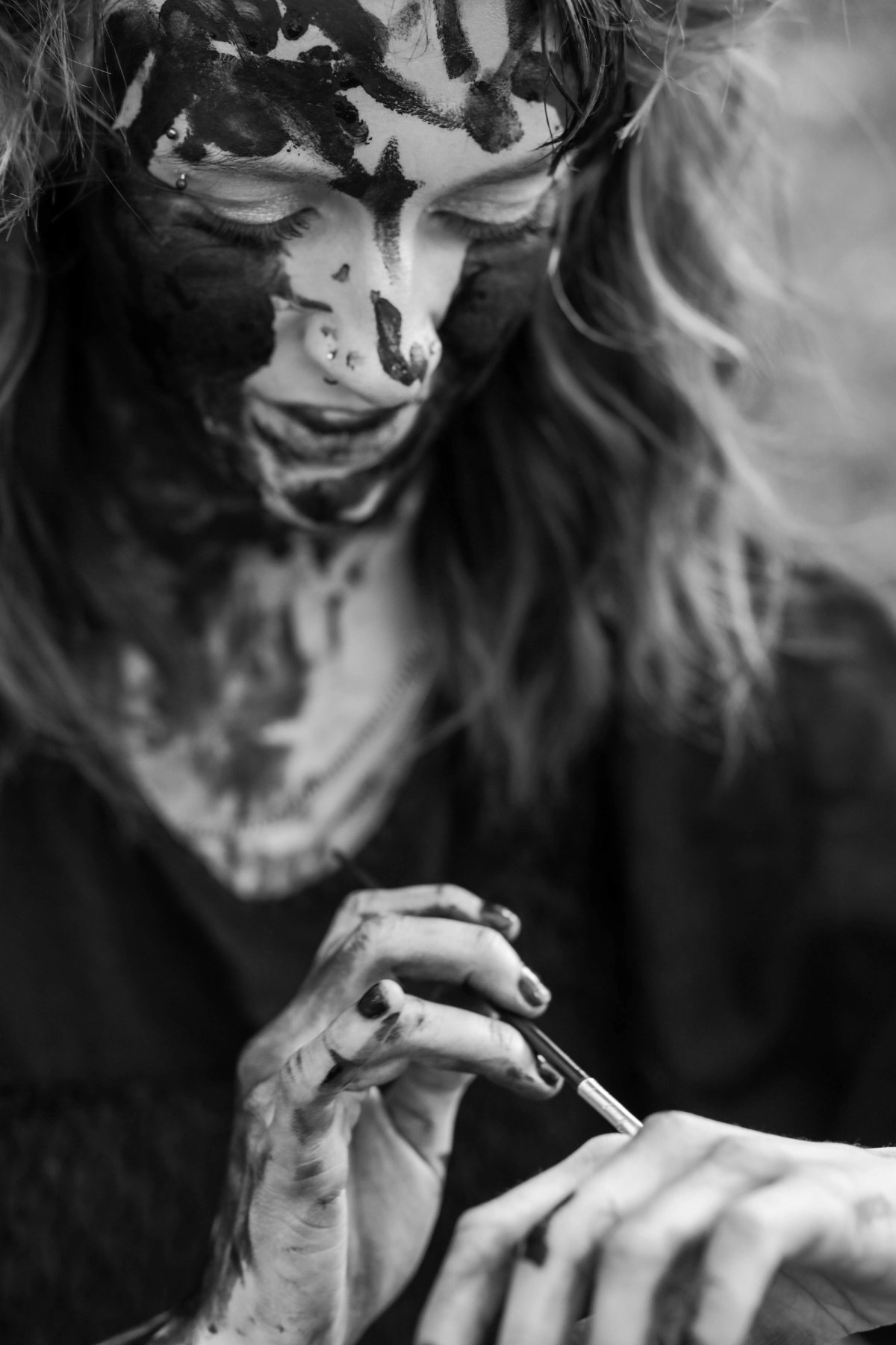 Looking down, eyes nearly closed, a woman with her face painted black dabs paint onto her own hand.