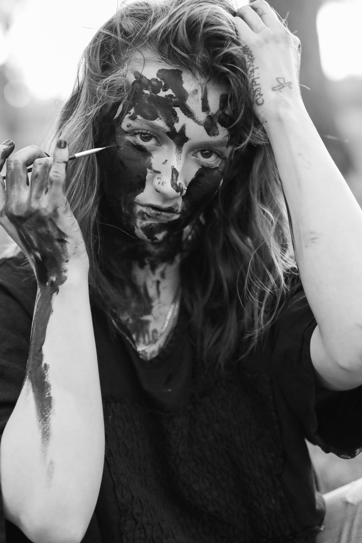 A woman with paint across her face holds a brush beside it, her other hand gripping her hair.