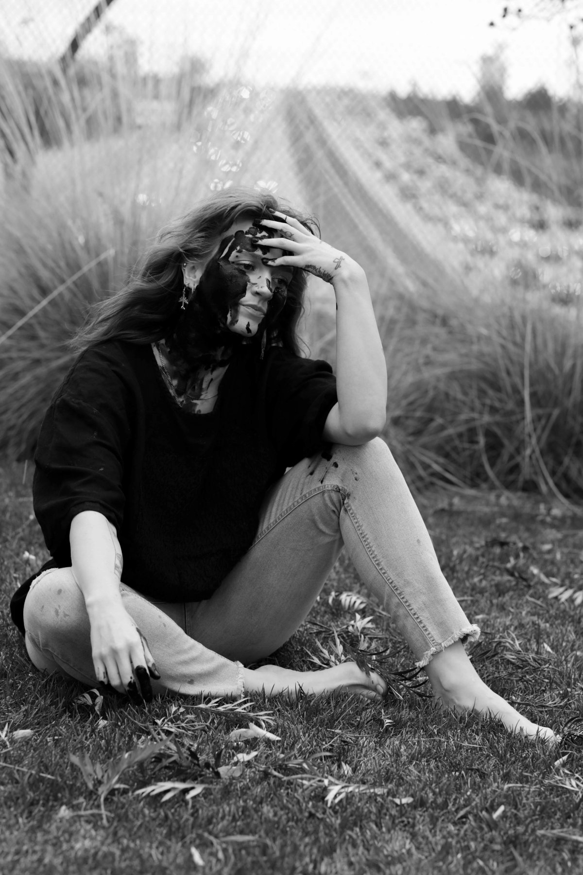 A woman sits in the grass by a fence, head in her hand, her face and neck covered in black paint.