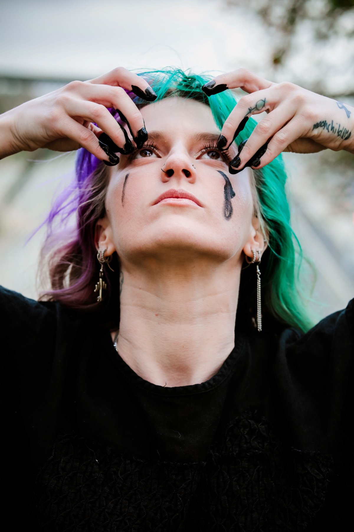 A woman with purple and green hair looks up, pressing black-painted fingertips around her eyes, paint running down one cheek.