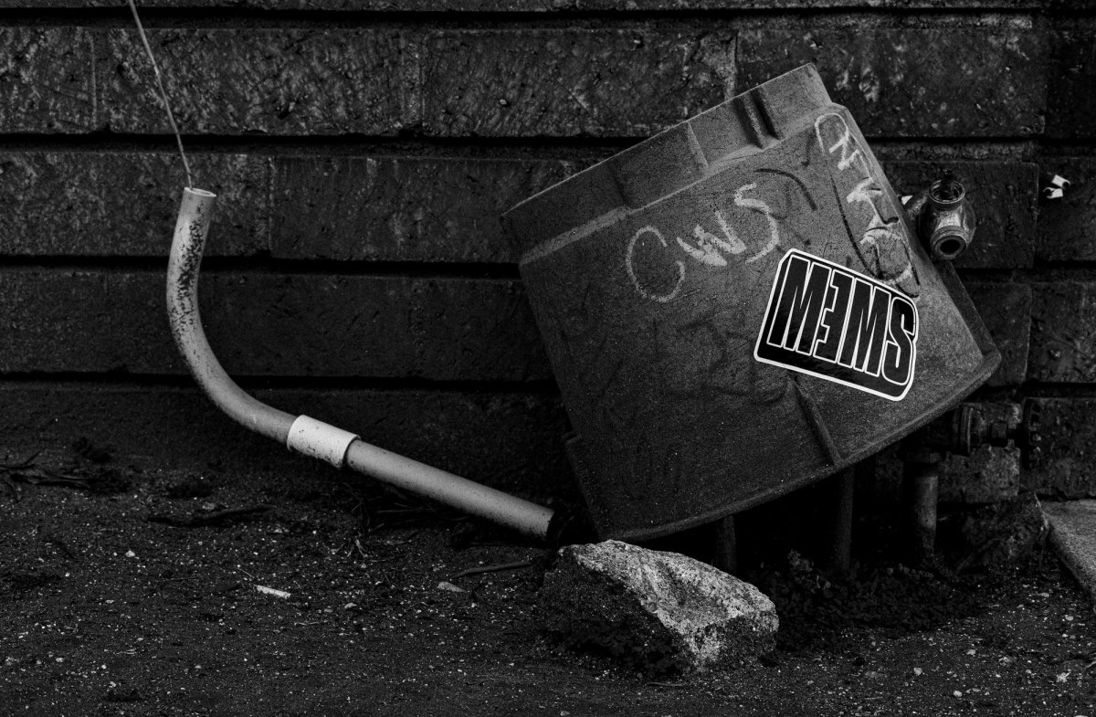 A dented gutter piece lies against a brick wall, tagged in chalk with a sticker stuck to it.