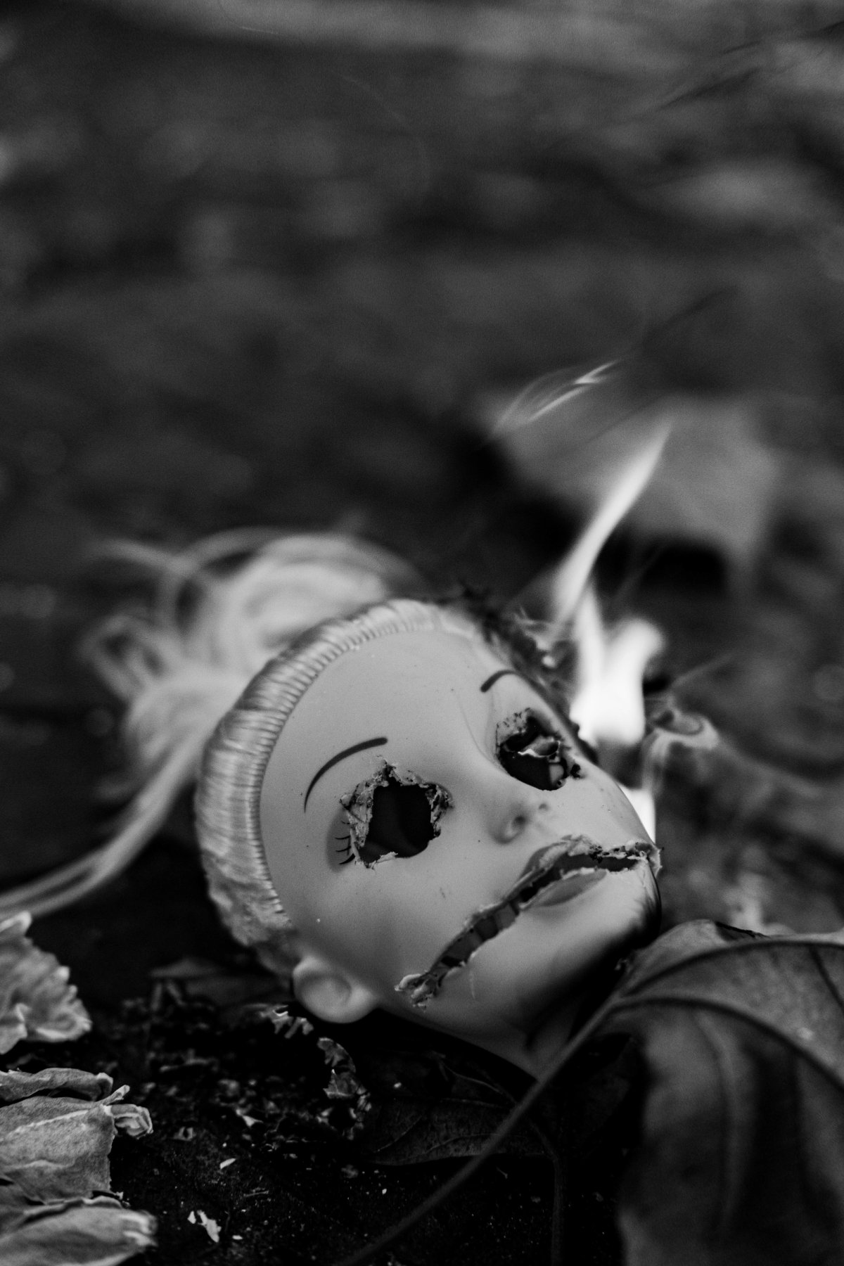 A doll's head lying in dark leaves, eyes and mouth burned open, a thin flame at its side.