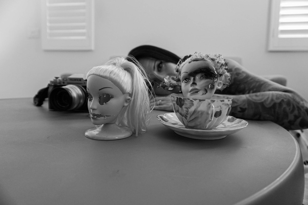 At eye level with the table: a camera, a doll head, and a scorched doll head inside a teacup, with a woman resting her head behind them.