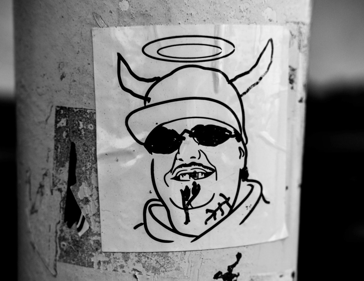 A hand-drawn sticker of a grinning man in a cap and sunglasses with devil horns and a halo.