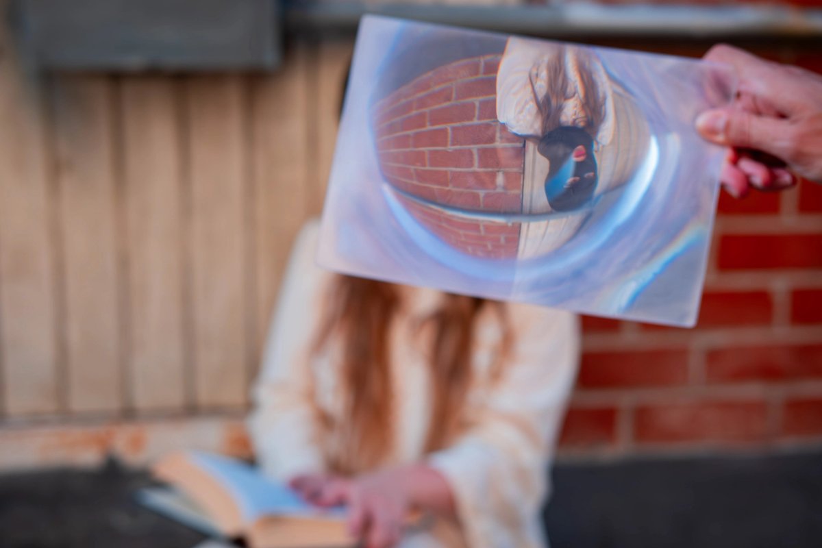 A hand holds a magnifying lens up, showing the masked woman upside down inside it.