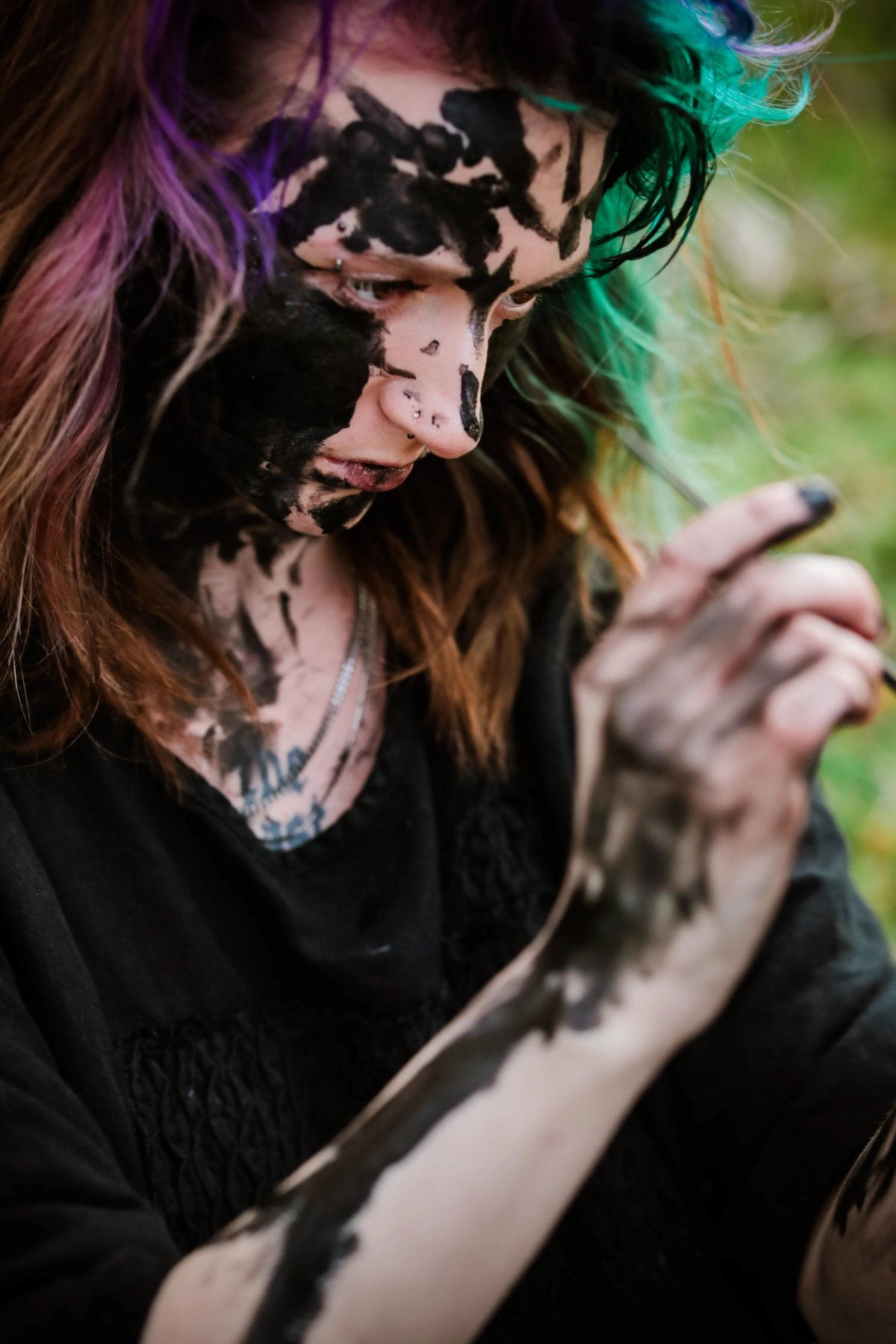 Looking down, a woman with purple and green hair, her face covered in black paint, smears paint along her arm.
