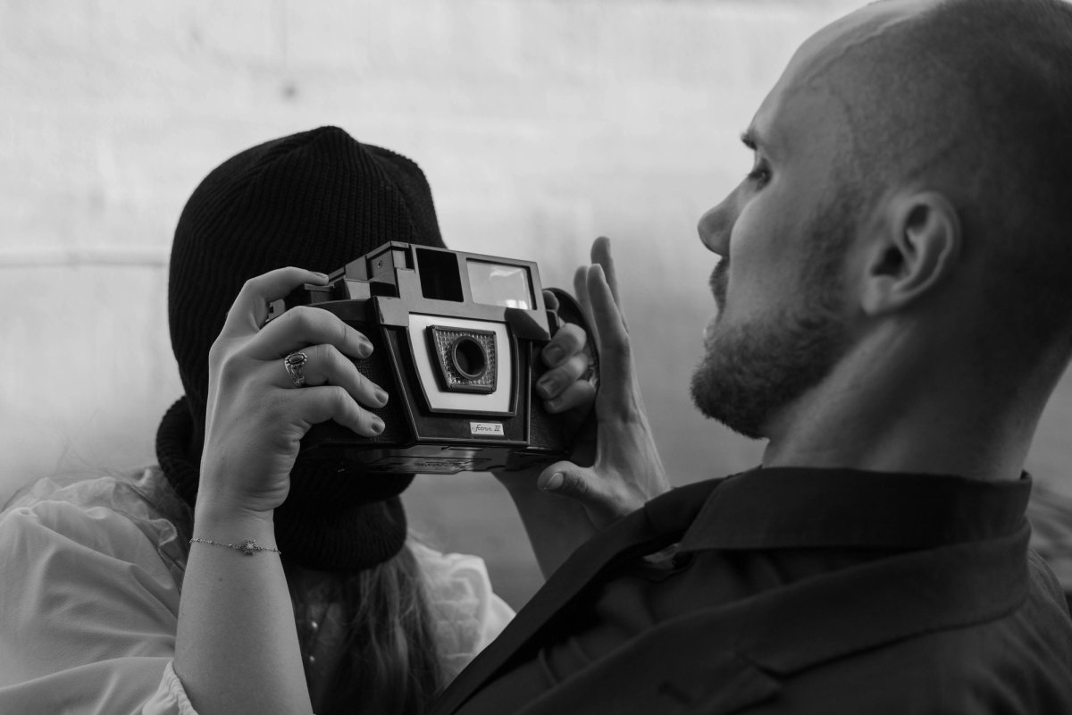 The masked woman raises an old camera to photograph the man beside her, who looks away.