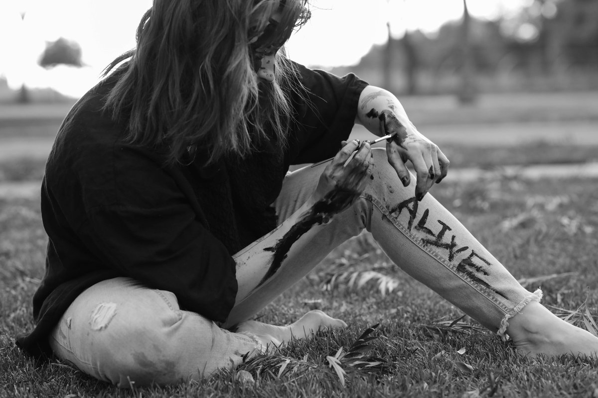 A woman sitting in the grass paints the word 'ALIVE' down the leg of her jeans.