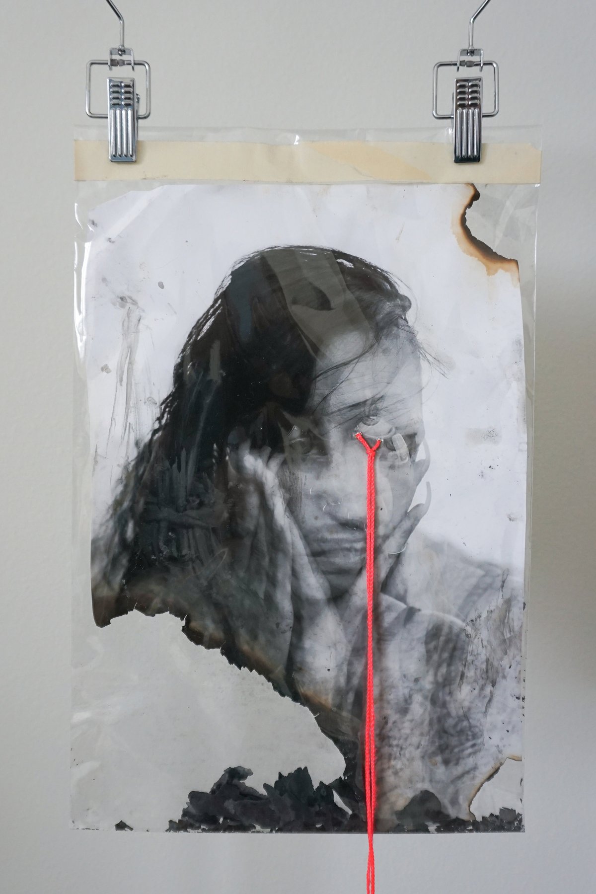A burned print of a woman's face, clipped up on a wall, a red string running down from her eye.