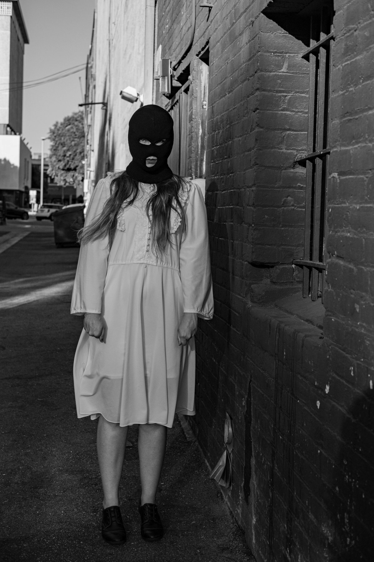 A masked woman in a white dress stands still at the end of a narrow brick alley.