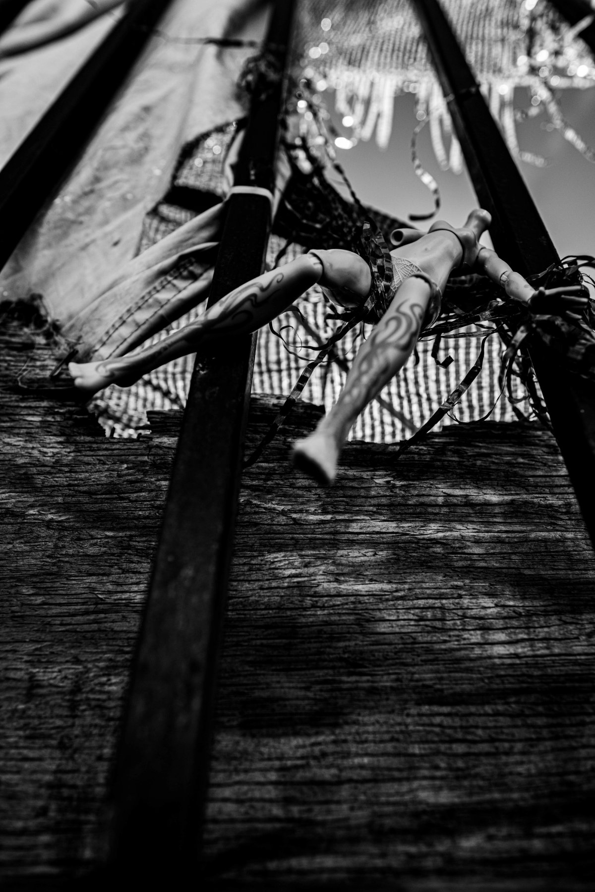 A doll tied with tinsel and ribbon hangs between two black bars, legs splayed.