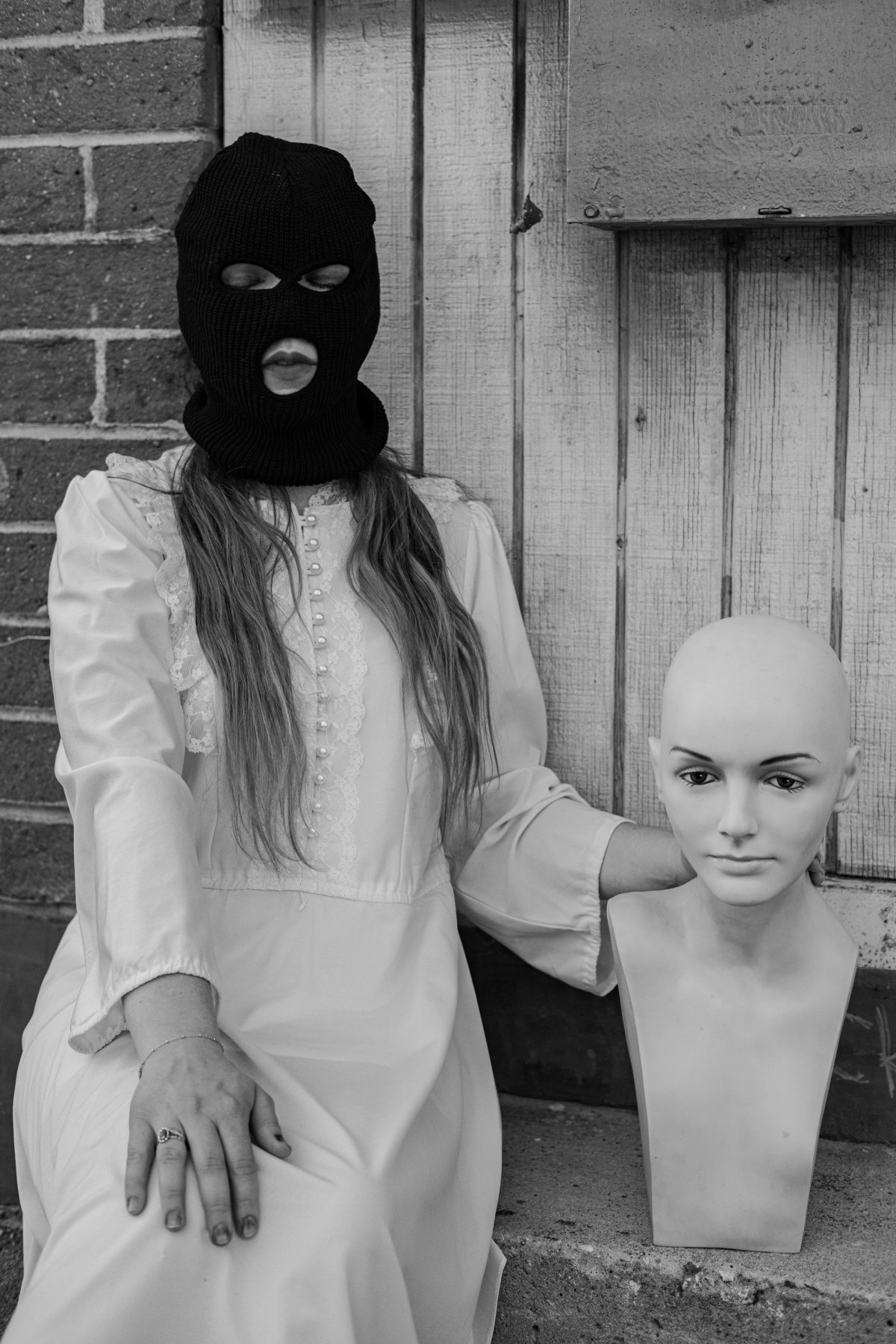 A masked woman in white sits beside a bald mannequin head, her hand resting on it.