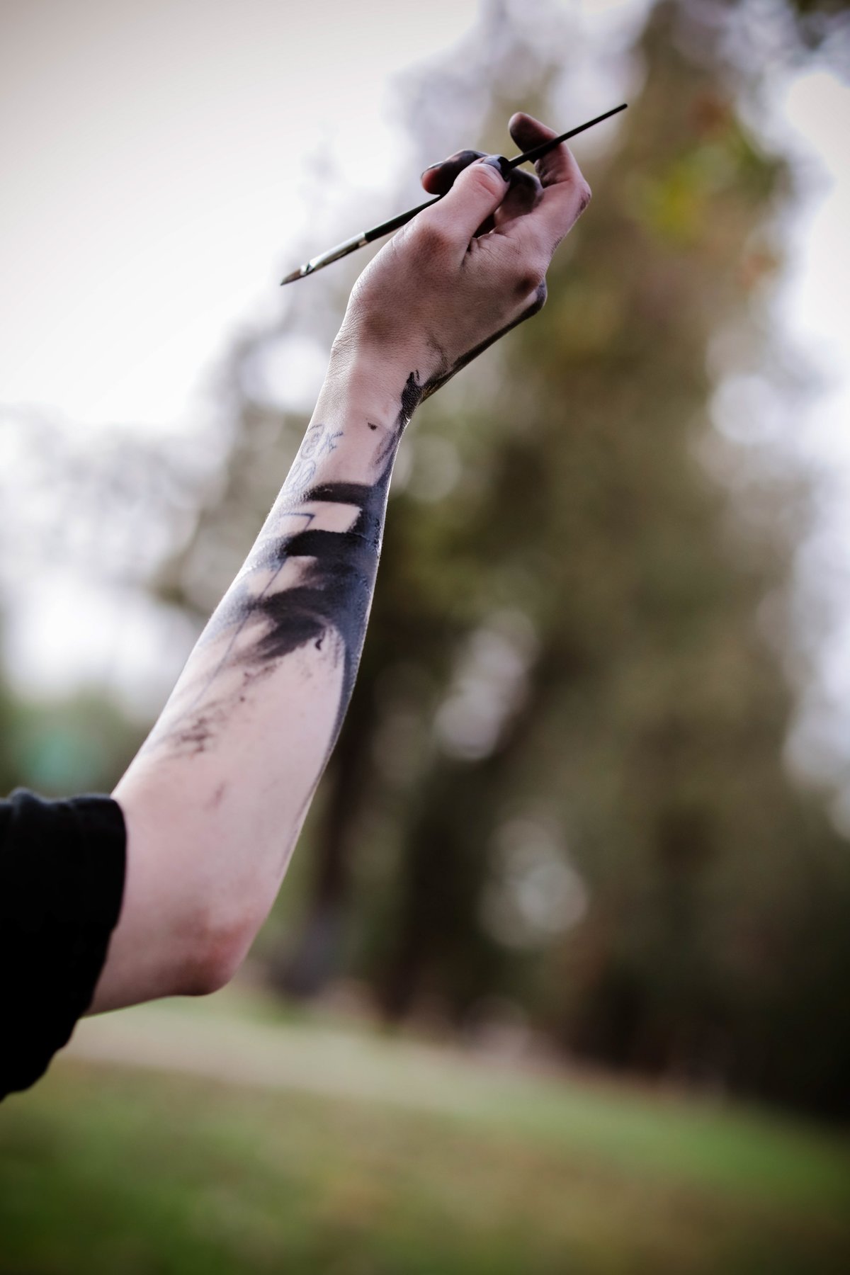 A single arm raised against blurred trees, holding a thin brush, the forearm brushed with black paint.