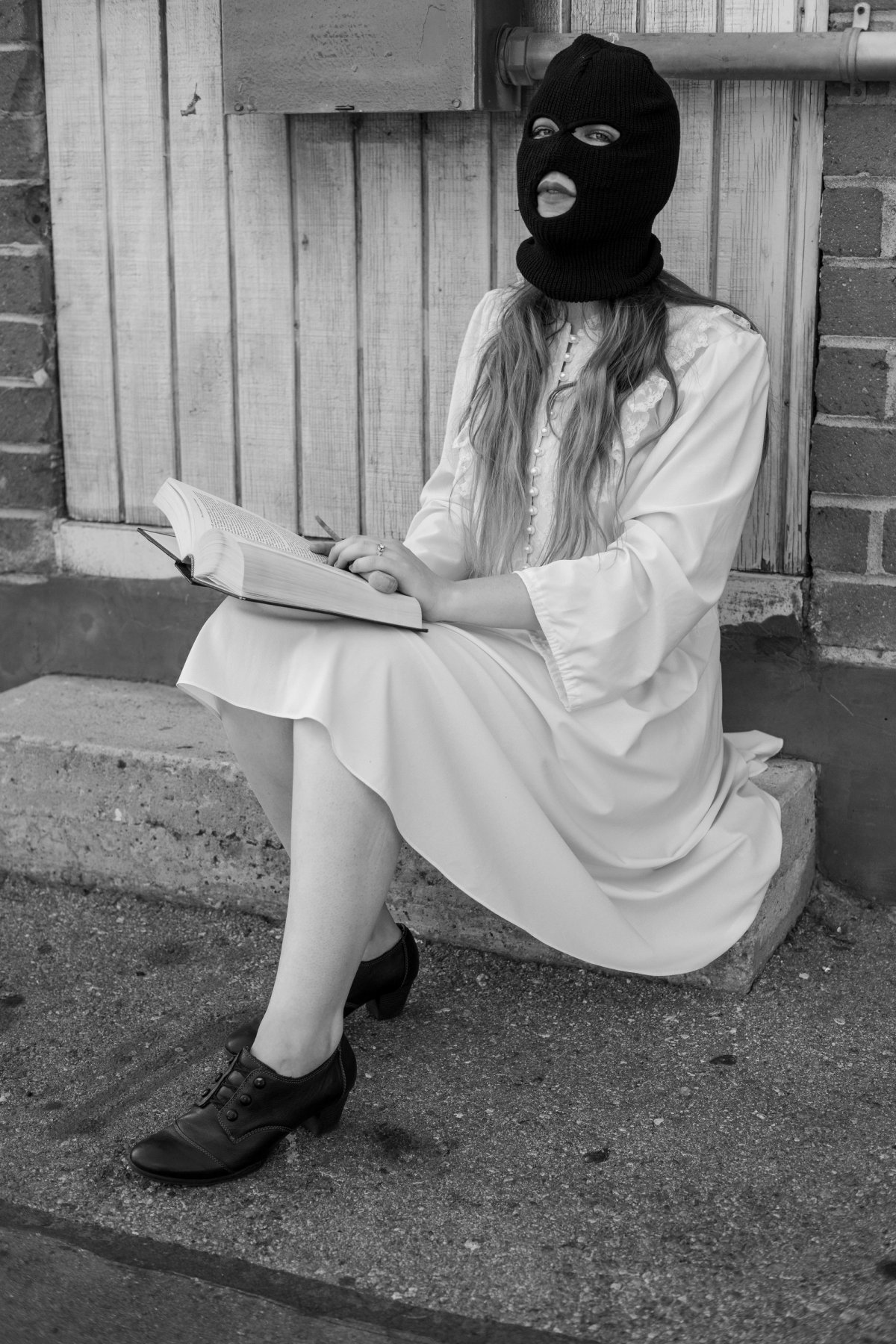 A woman in a black ski mask and white dress sits on a step with an open book.