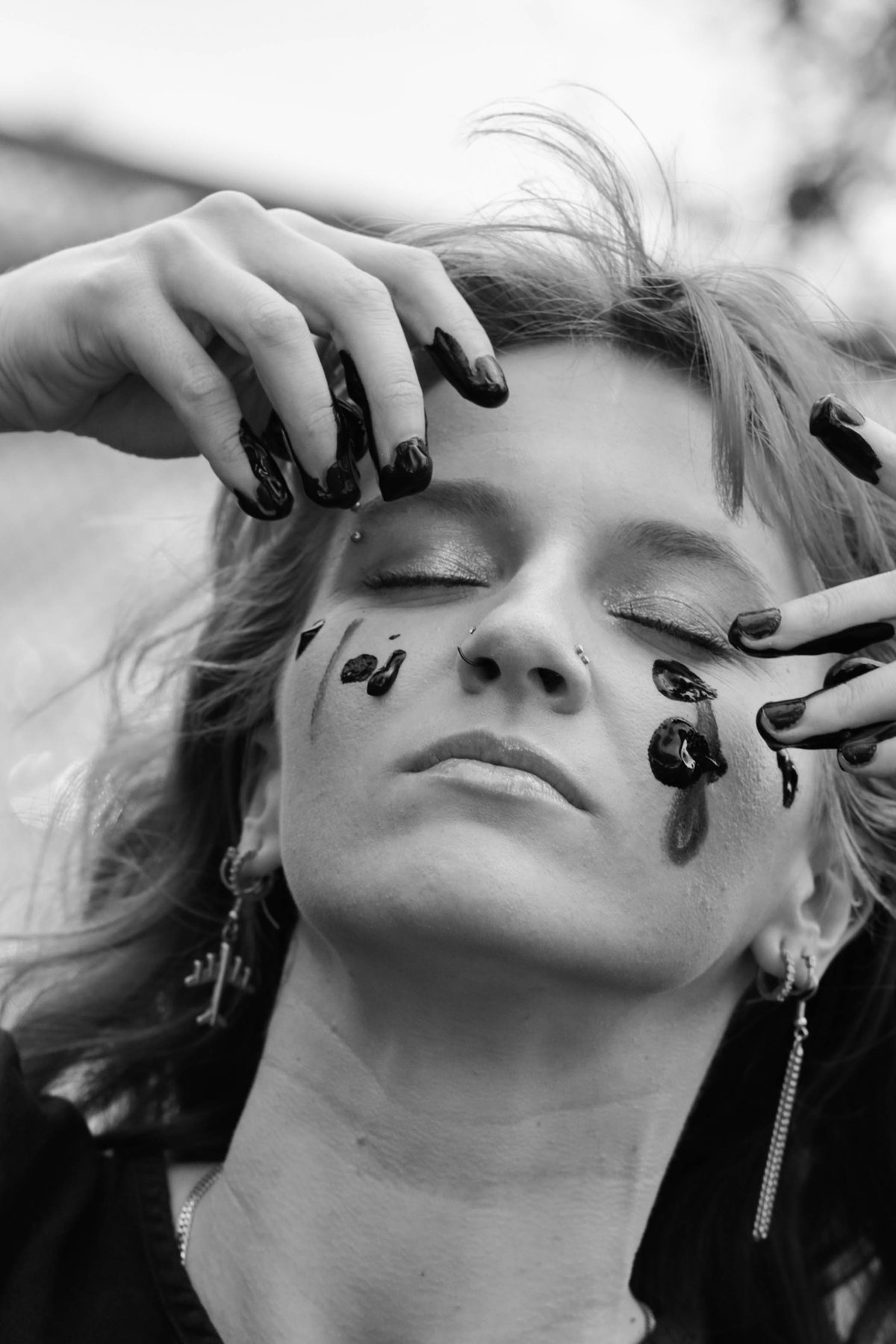 Eyes closed, face tilted up, a woman presses black-painted fingertips to her face, paint dripping down her cheek.