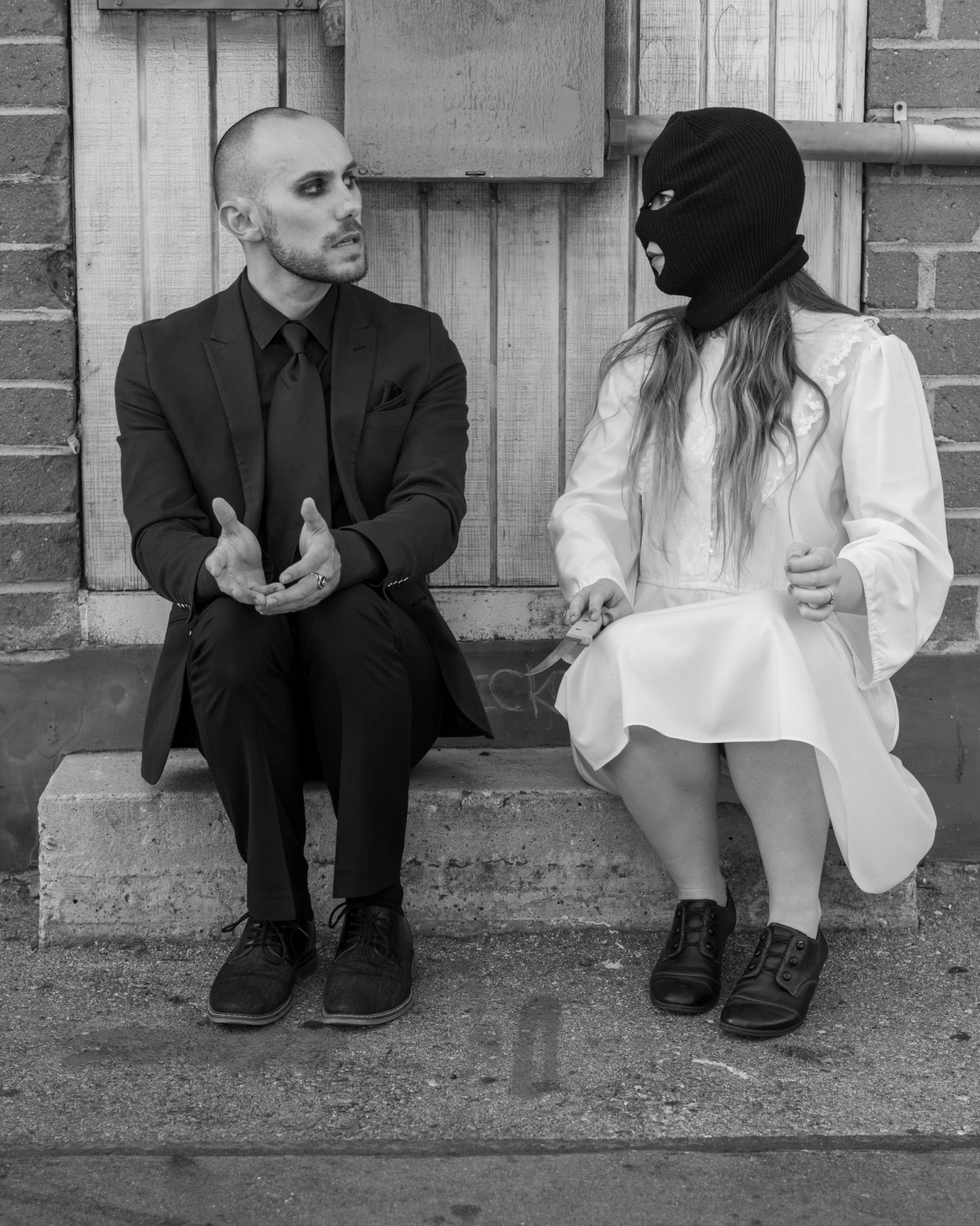 A man in a black suit and a masked woman in a white dress sit side by side on a step, talking.