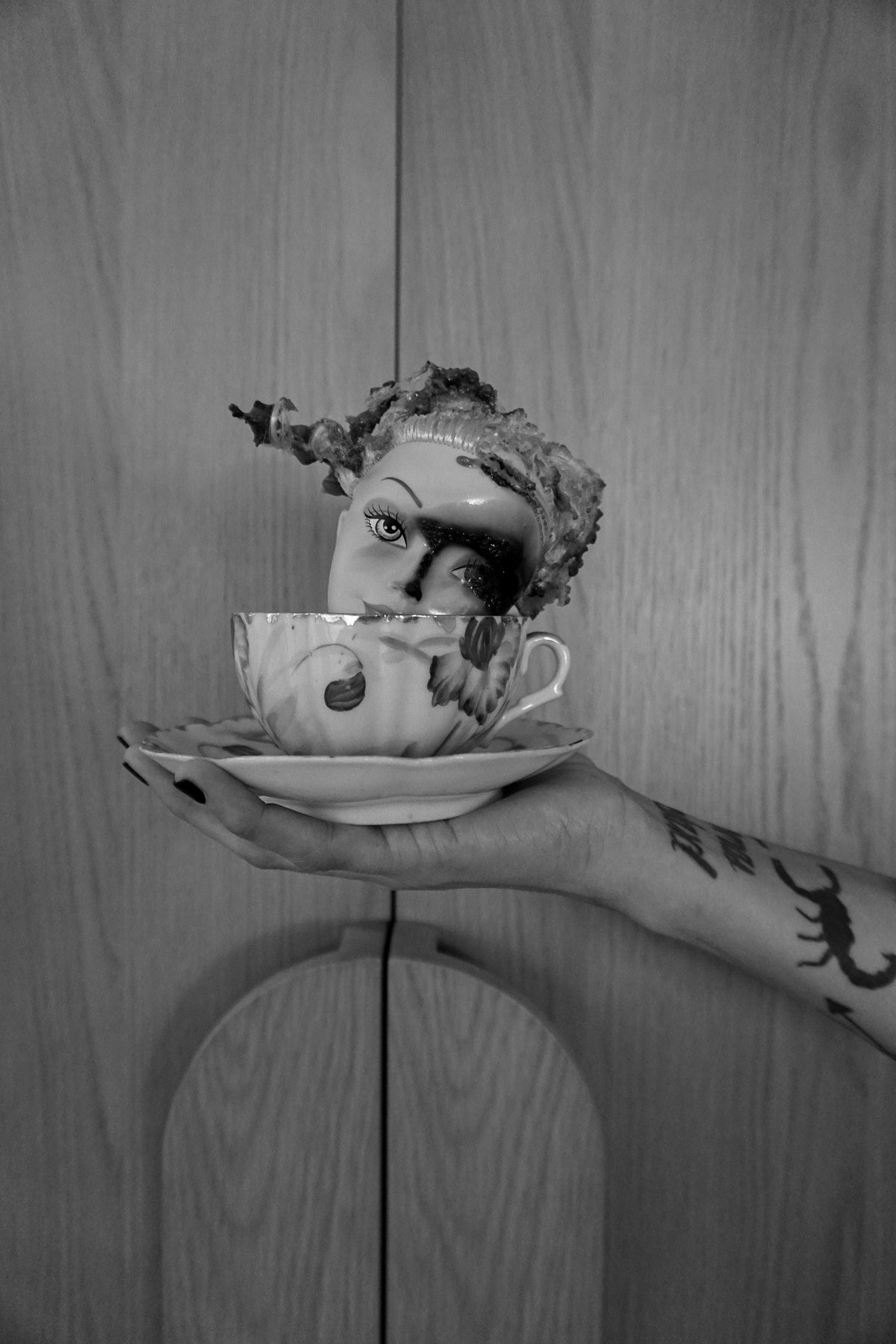 A tattooed hand holds out a teacup on a saucer, a doll head with a scorched eye inside it.
