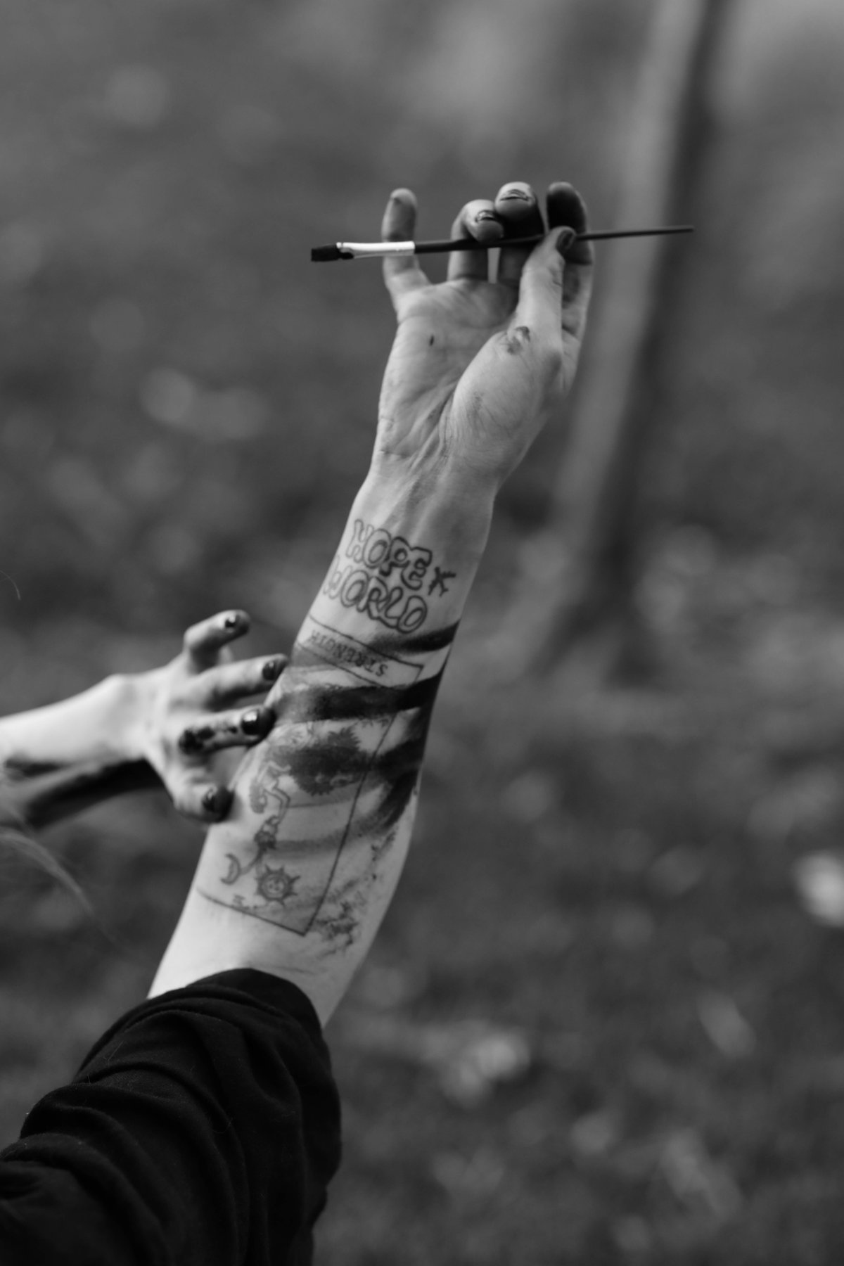 A tattooed forearm raised high, the hand holding a brush, another paint-stained hand reaching toward it.