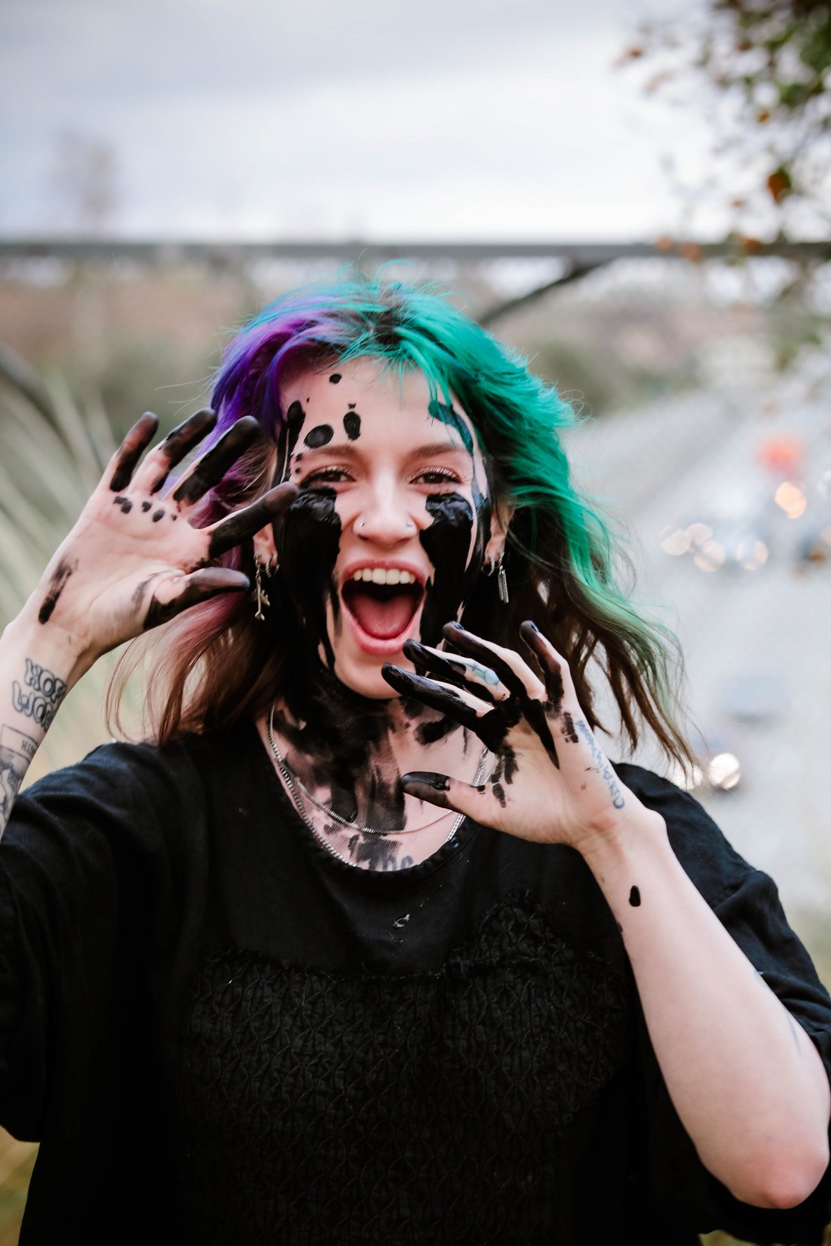 A woman with purple and green hair screams, laughing or crying out, her face and hands smeared with black paint.
