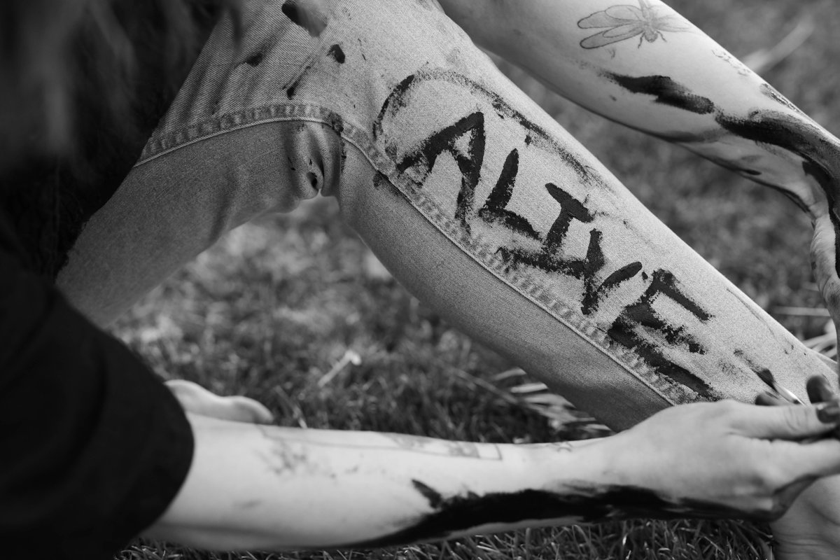 Close on a leg in light jeans with 'ALIVE' painted on it in black, hands and arms smeared with paint.
