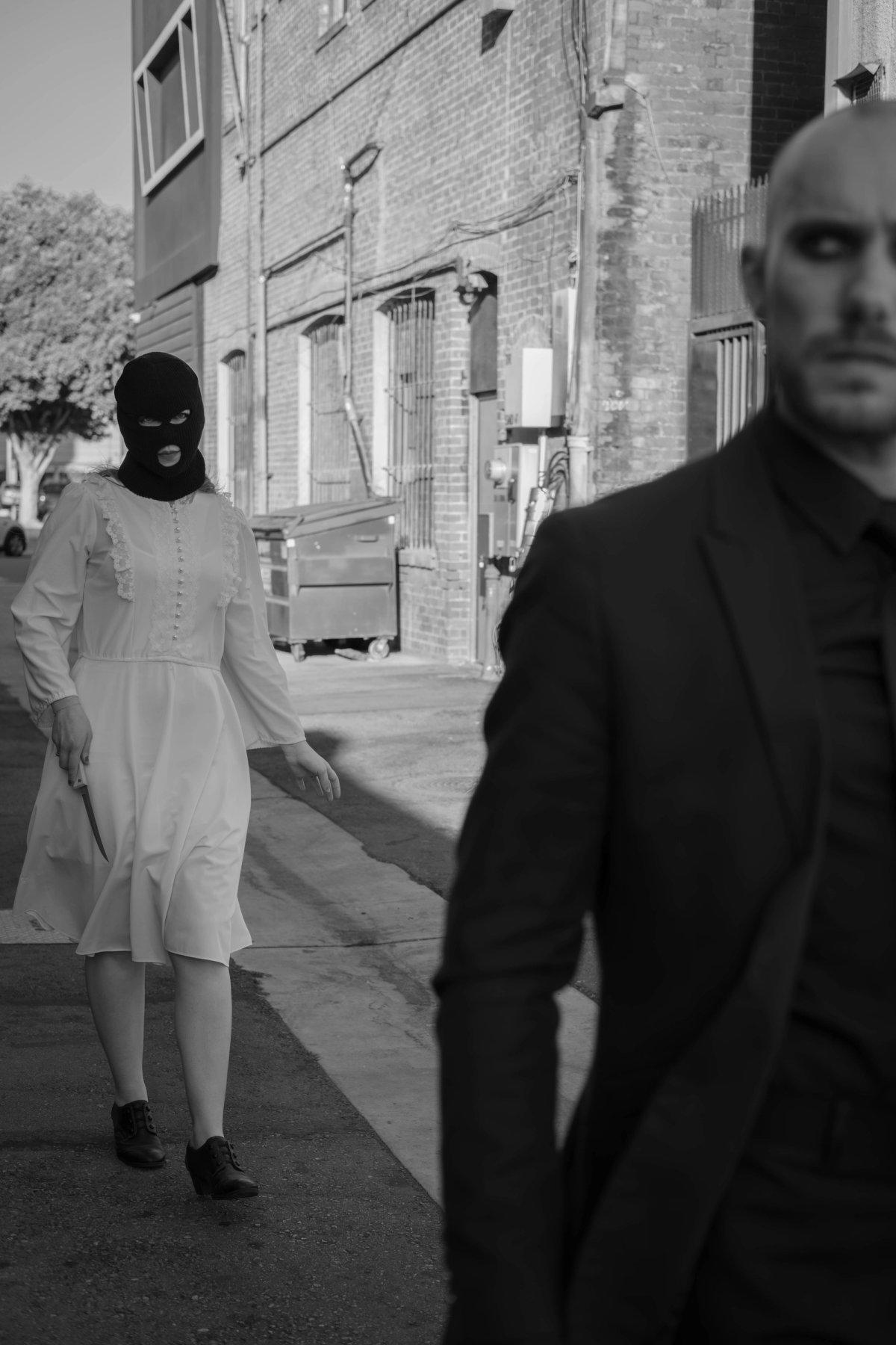 In an alley, a man in black looks back while a masked woman in white walks behind him holding a knife.