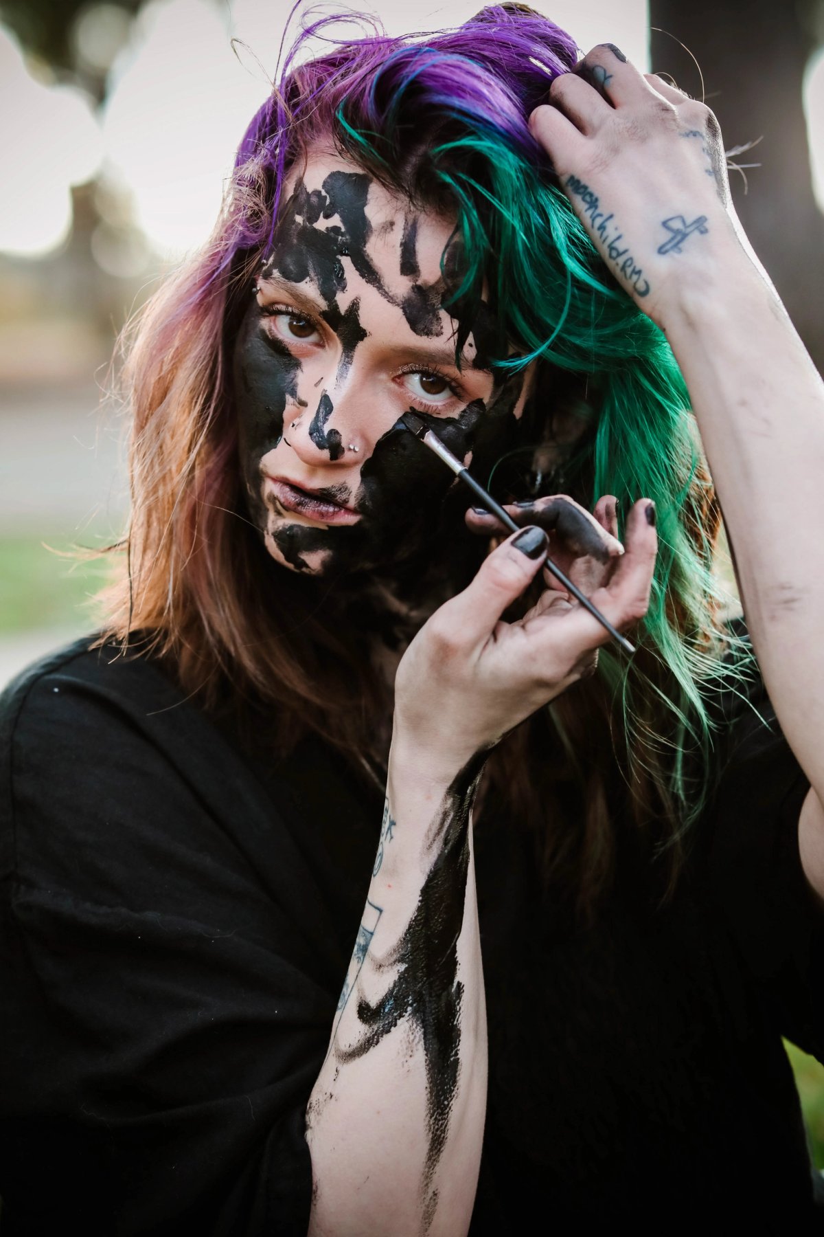 A woman with purple and green hair paints her own face black with a small brush.