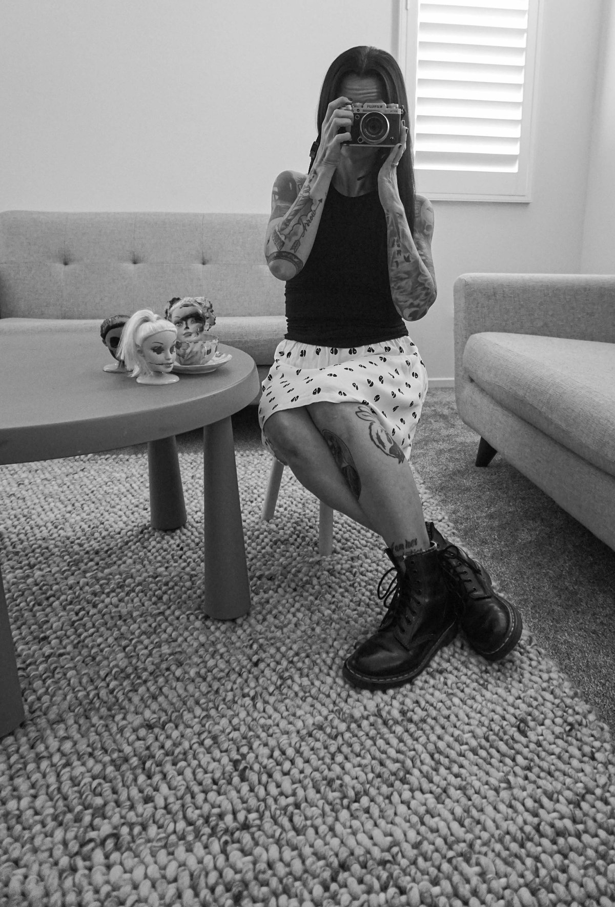 A woman sits at a small table photographing the viewer, doll heads and a teacup beside her.