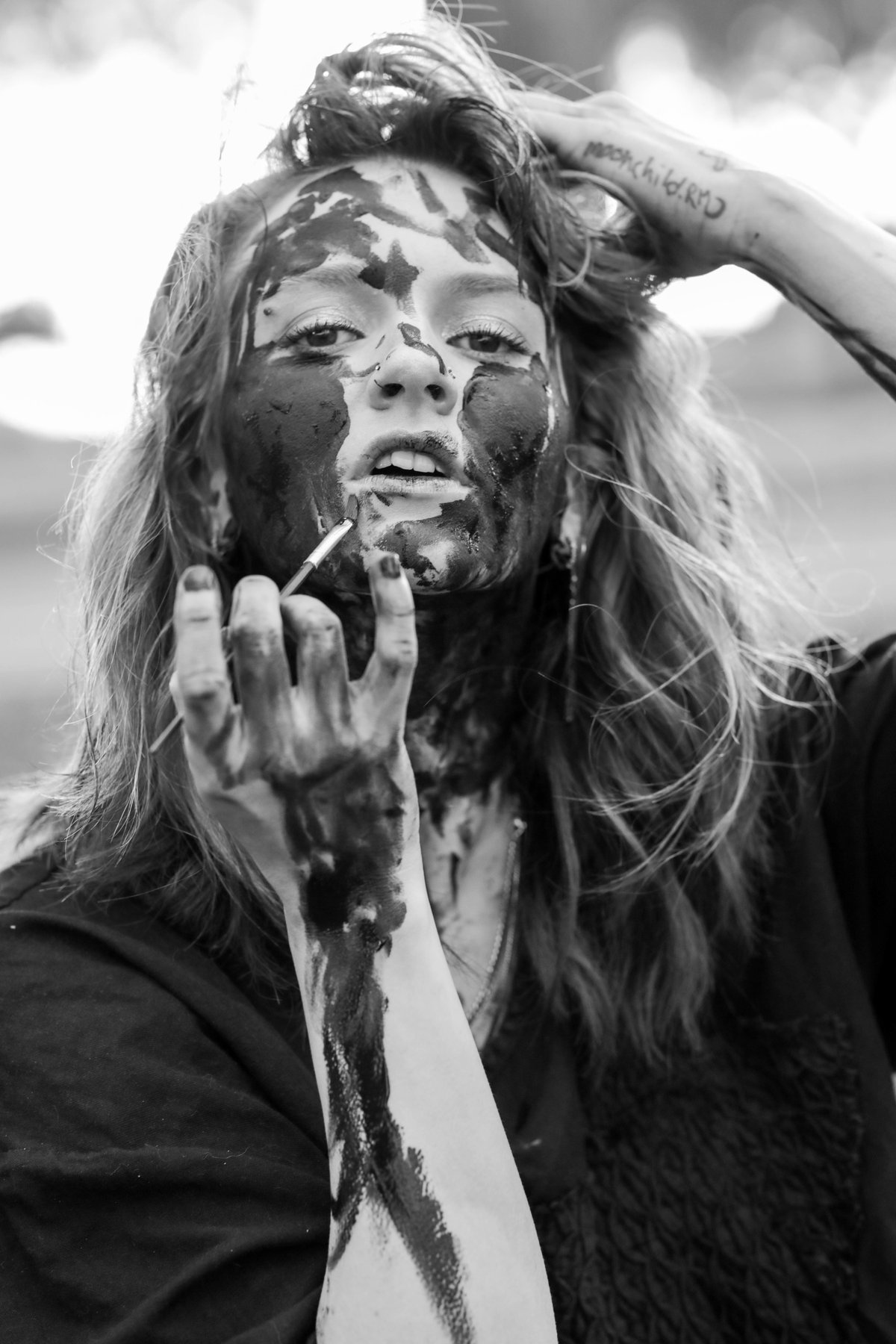 A woman with her face painted black brings a brush to her lips, her other hand in her hair.