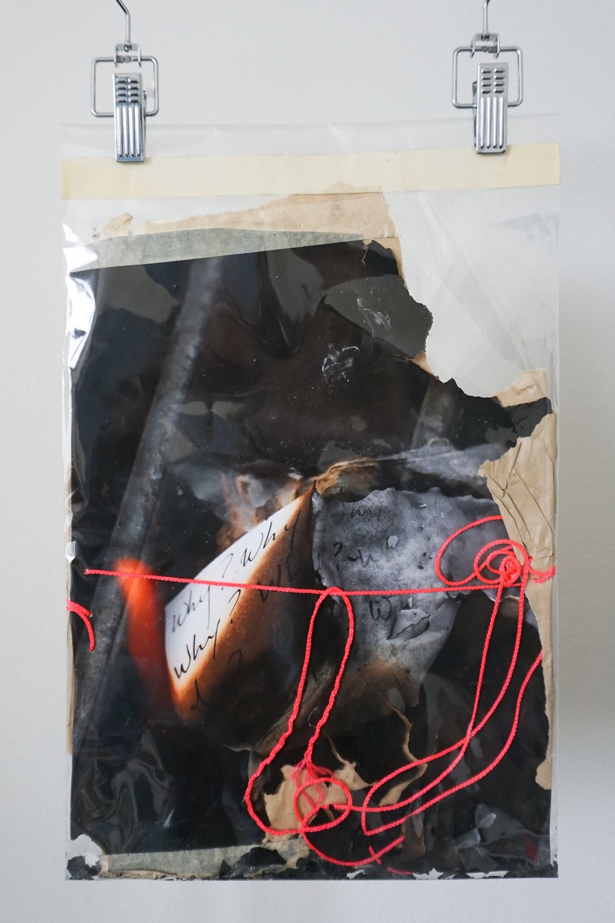 A burned collage clipped to a wall: a curl of 'Why?' paper caught in flame, red string tied across it.