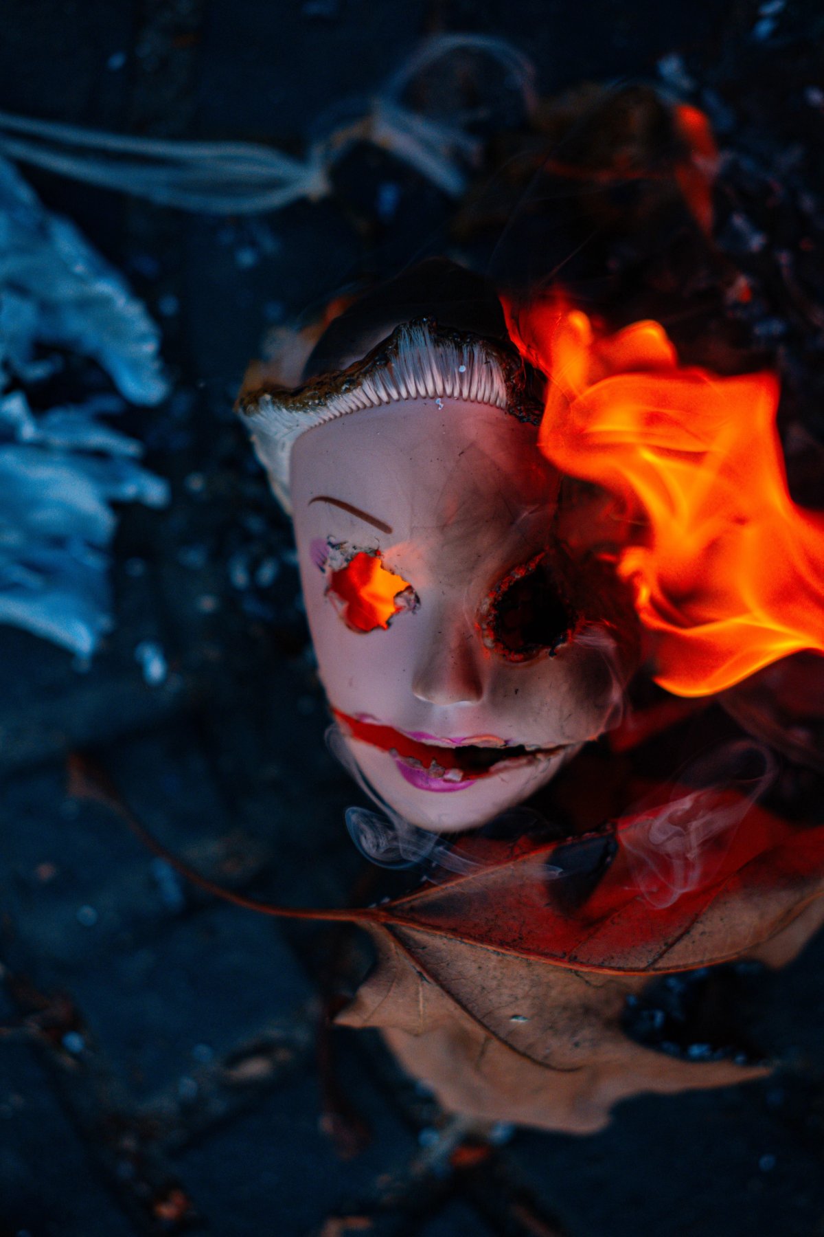 A doll's head on fire, one eye glowing orange, smoke curling over a fallen leaf.