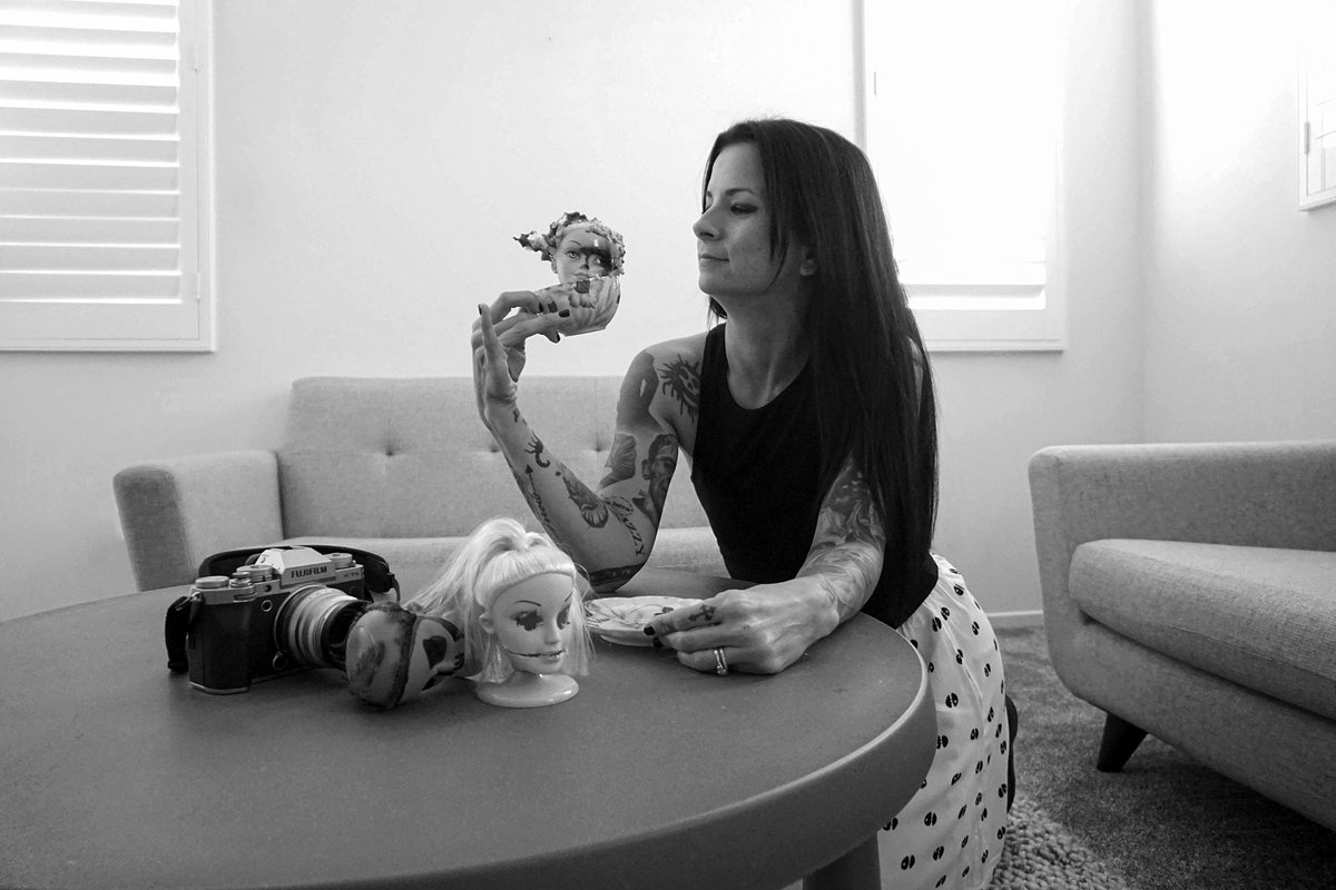 A woman at the table raises the teacup with the doll head inside, beside a camera.