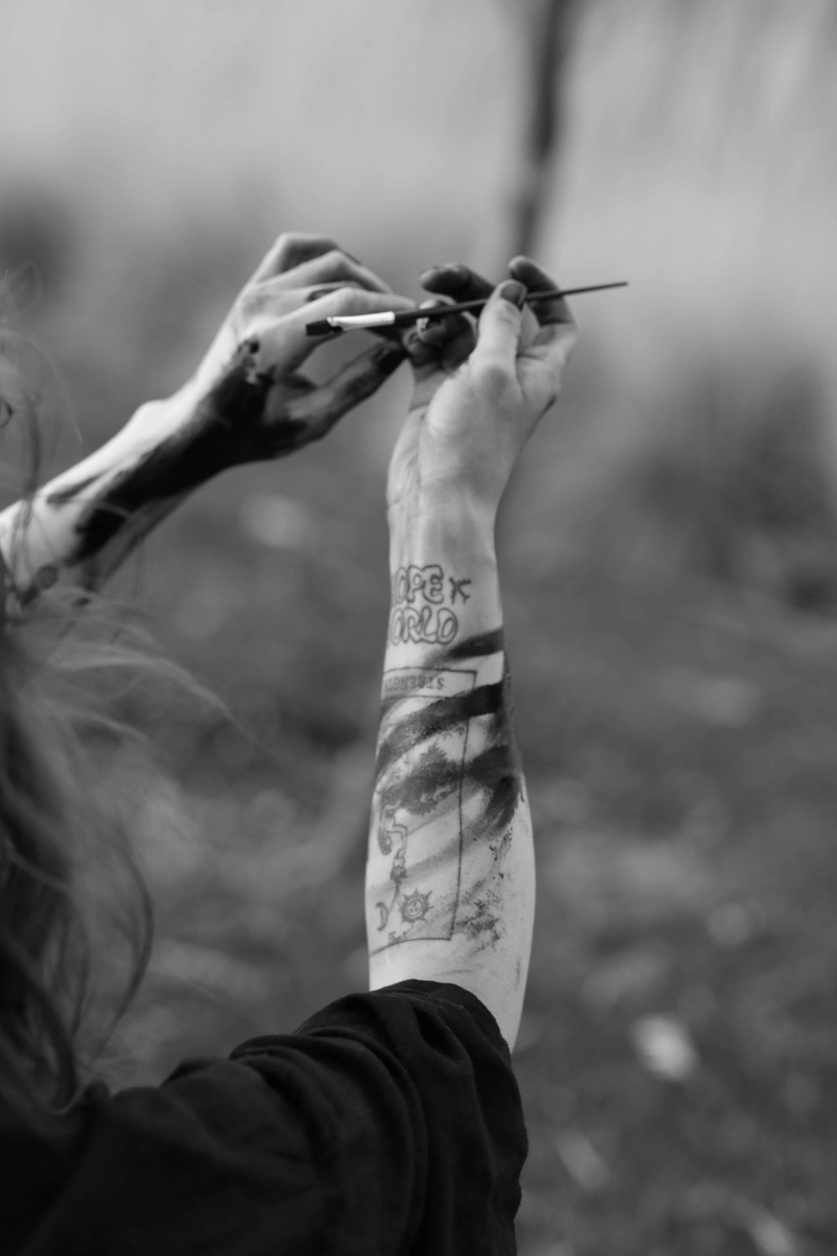 Two hands raised together, passing a brush, a tattooed forearm streaked with black paint.
