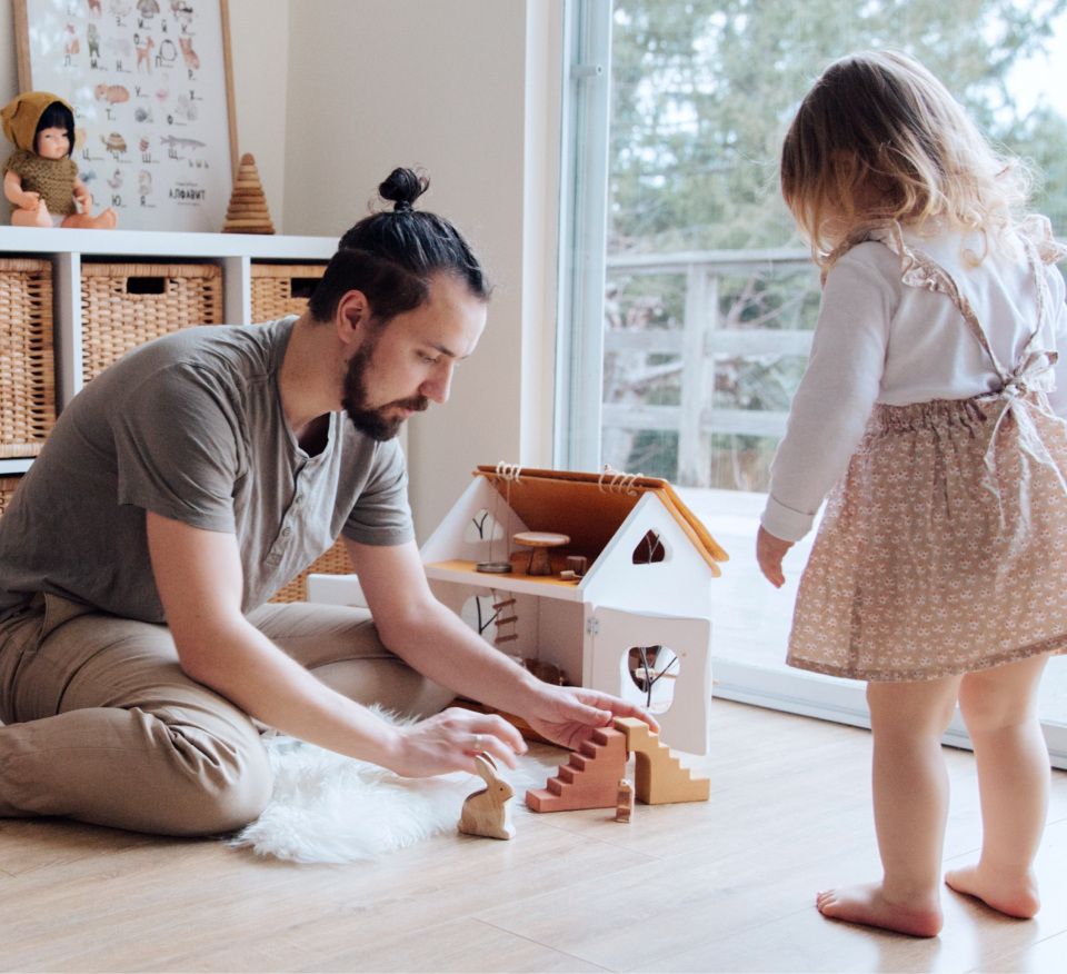 Man with manbun sitting on the floor using toys to play with a child.