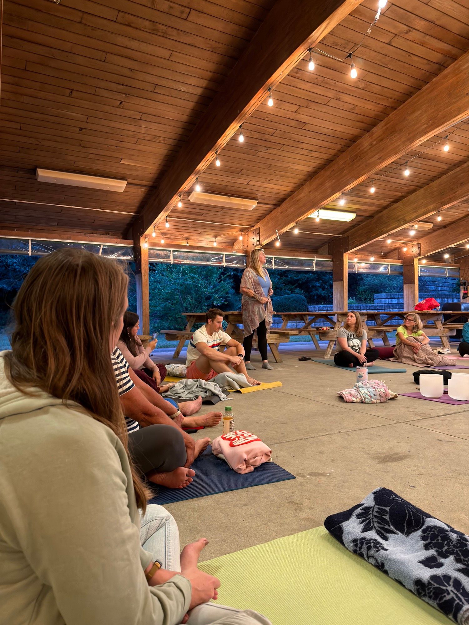 Attendees seated on mats listening, seen from behind, under the pavilion’s string lights.