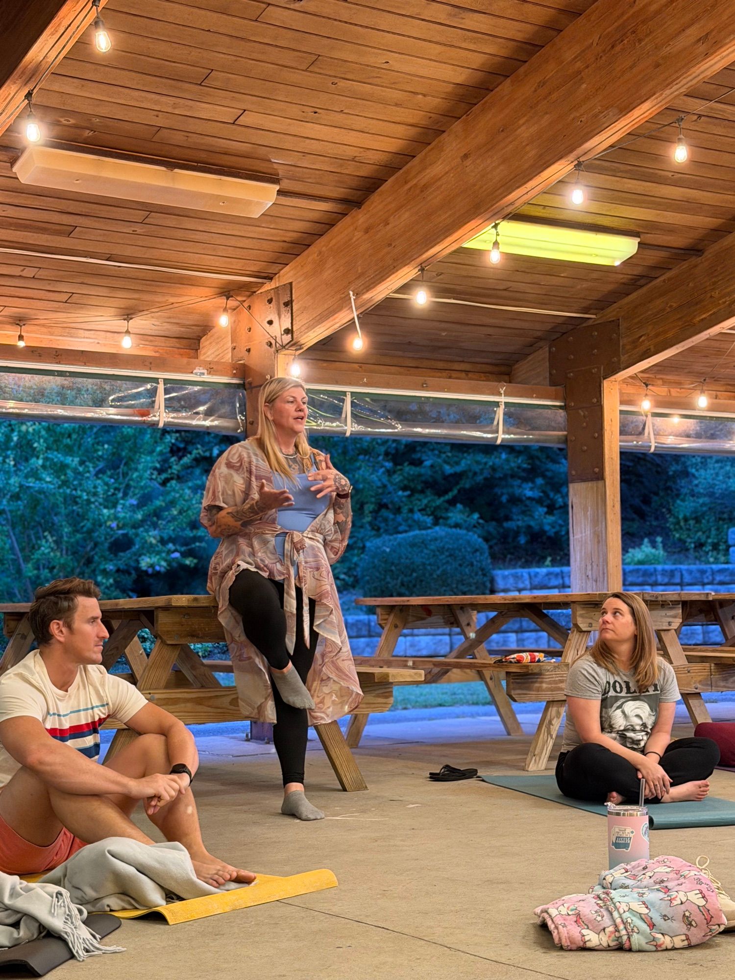 A woman standing and speaking to the group, who are seated on mats around her.
